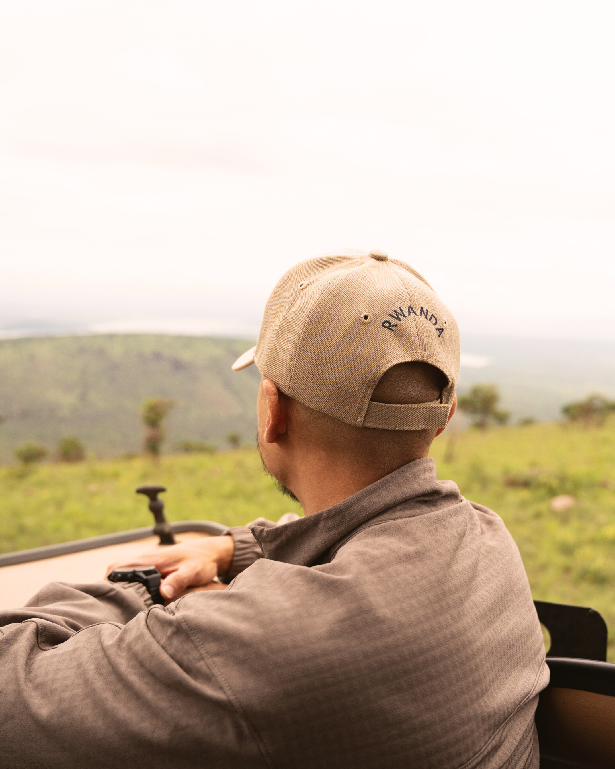 Person driving a safari vehicle in Rwanda, wearing a beige cap with 'RWANDA' embroidered, overlooking a lush, green landscape with hills and scattered trees.