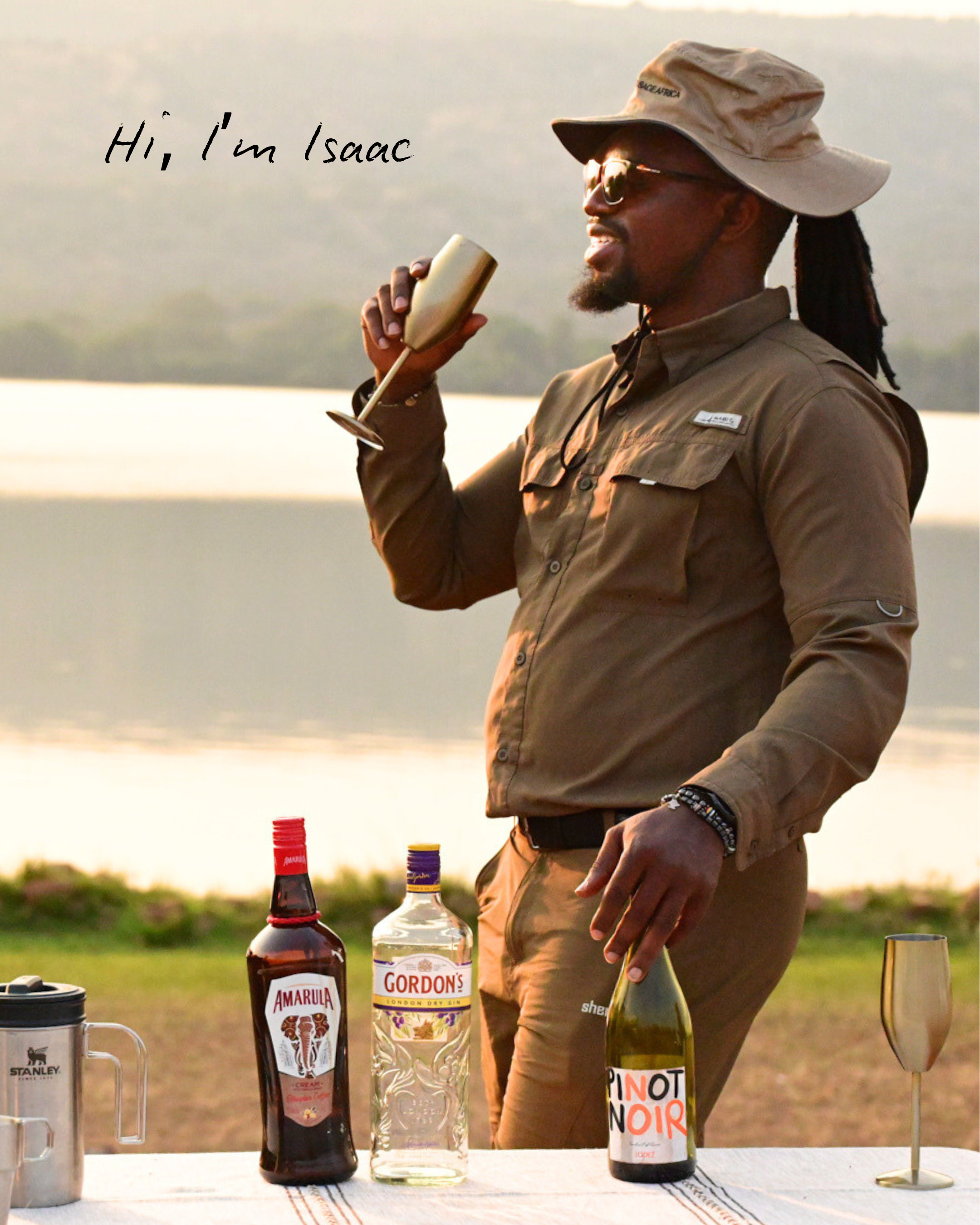 A man with dreadlocks, wearing sunglasses, a beige hat, and a brown outdoor shirt, stands outdoors near a body of water, holding a champagne flute. On the table in front of him are bottles of Amarula, Gordon's gin, and Pinot Noir wine, along with a metal Mug and a gold-colored champagne flute.