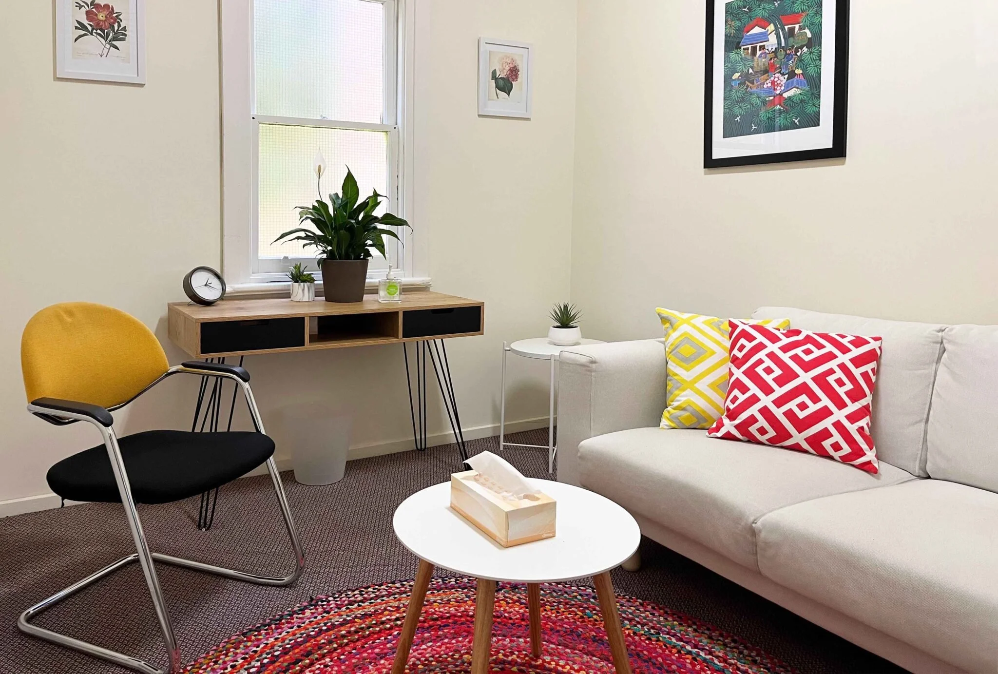 A cosy counselling office with a white sofa, additional chair, circular table and desk. There is art on the walls and a window.