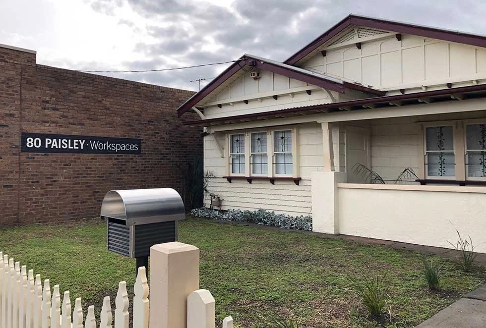 A small white house with a porch, next to a brick wall with a sign that reads '80 Paisley Workspaces.' There is a mailbox and some grass in the front yard.