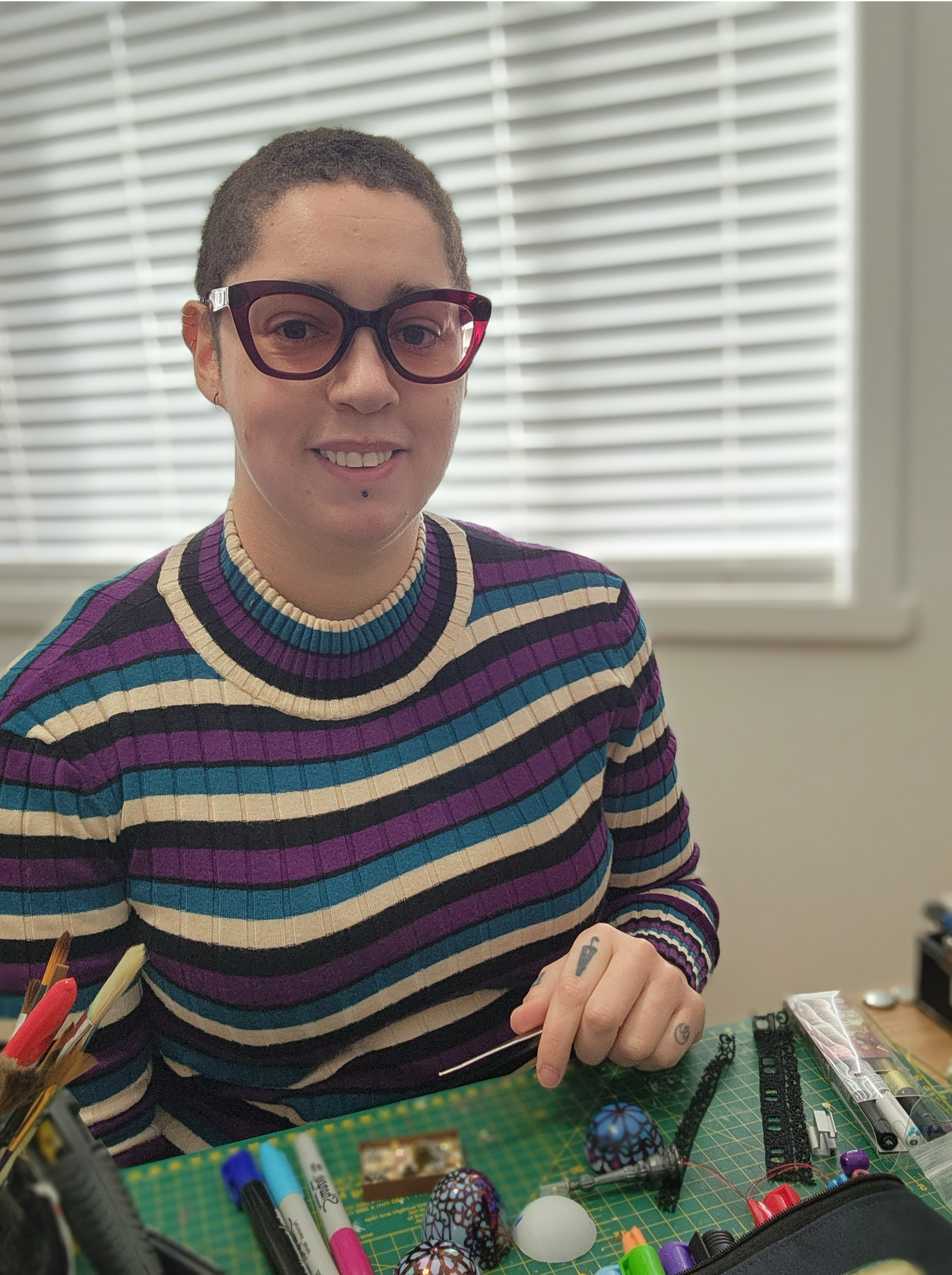 A person with short hair, wearing glasses and a striped sweater, sitting at a craft table with various crafting supplies and decorated eggs.