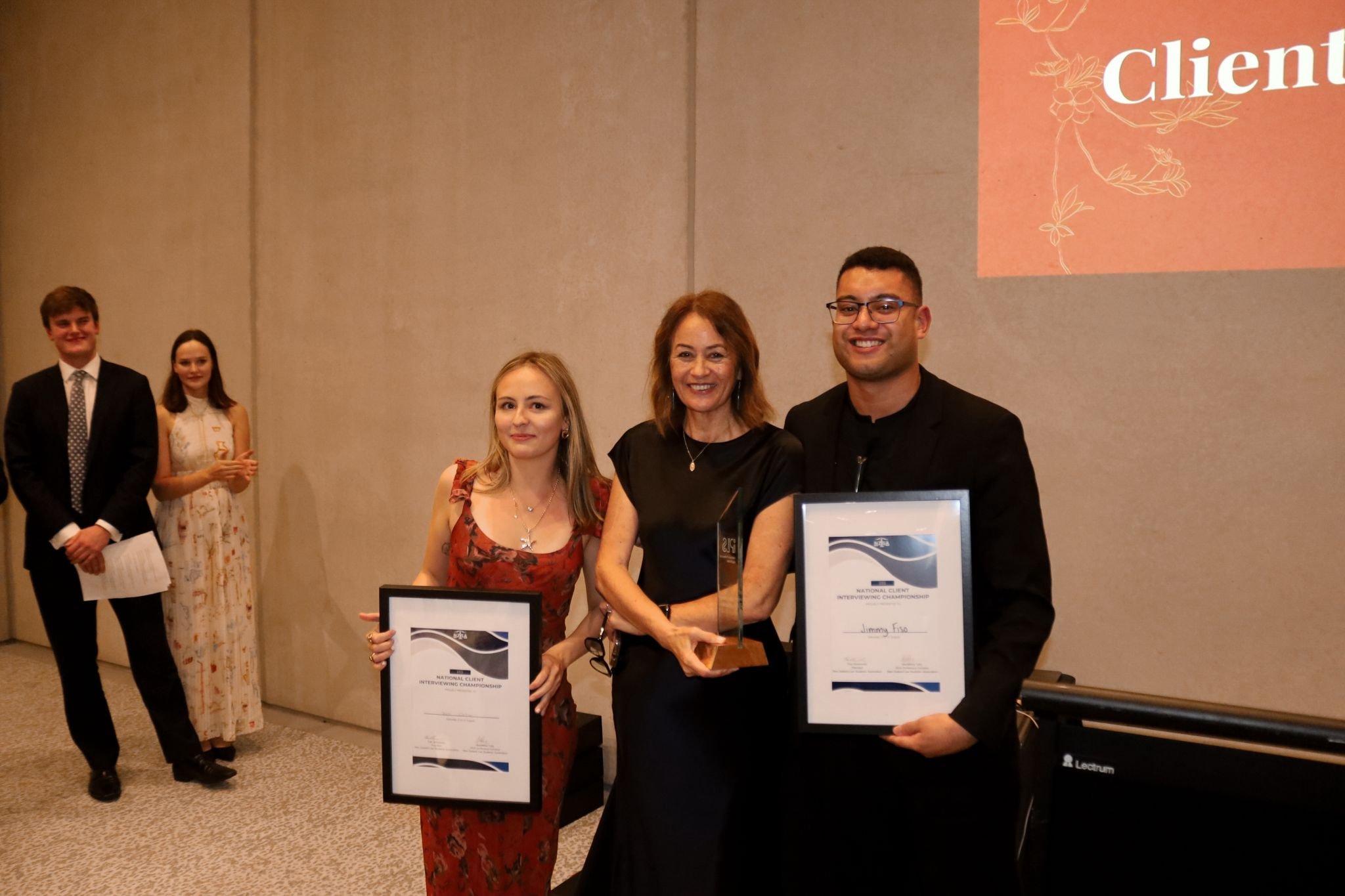 Group of people at an awards event; two individuals hold certificates, one holds a trophy. Other attendees stand in the background. Formal setting with a presentation projected on the wall.