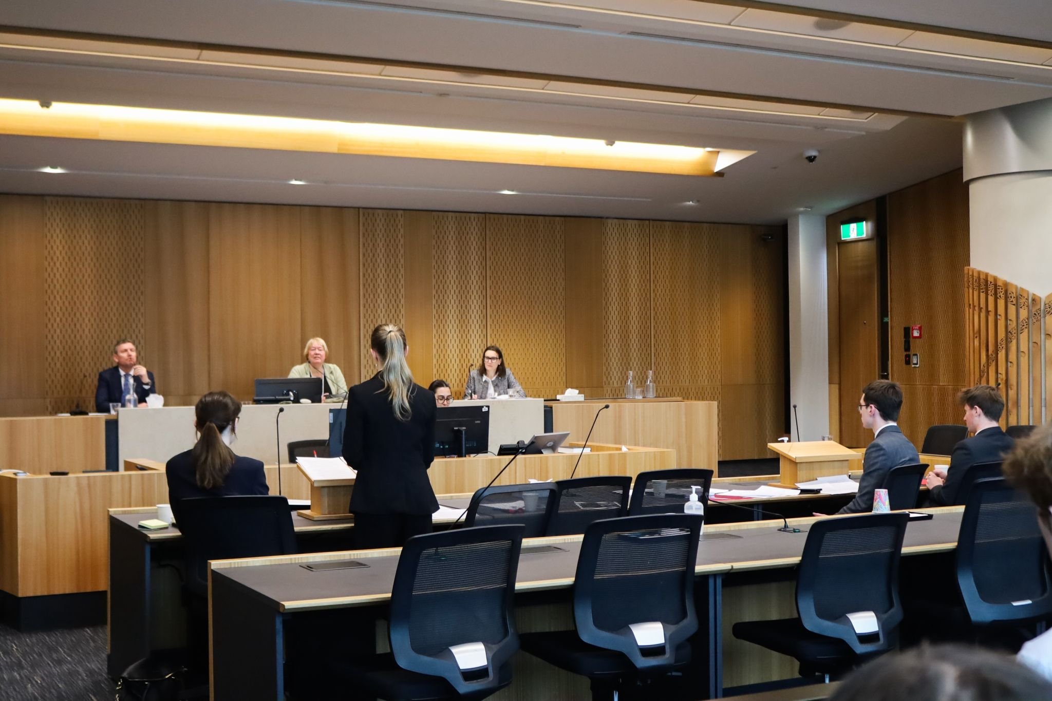 A courtroom scene with people seated at desks, a woman standing, and a panel of judges or officials at a higher desk.