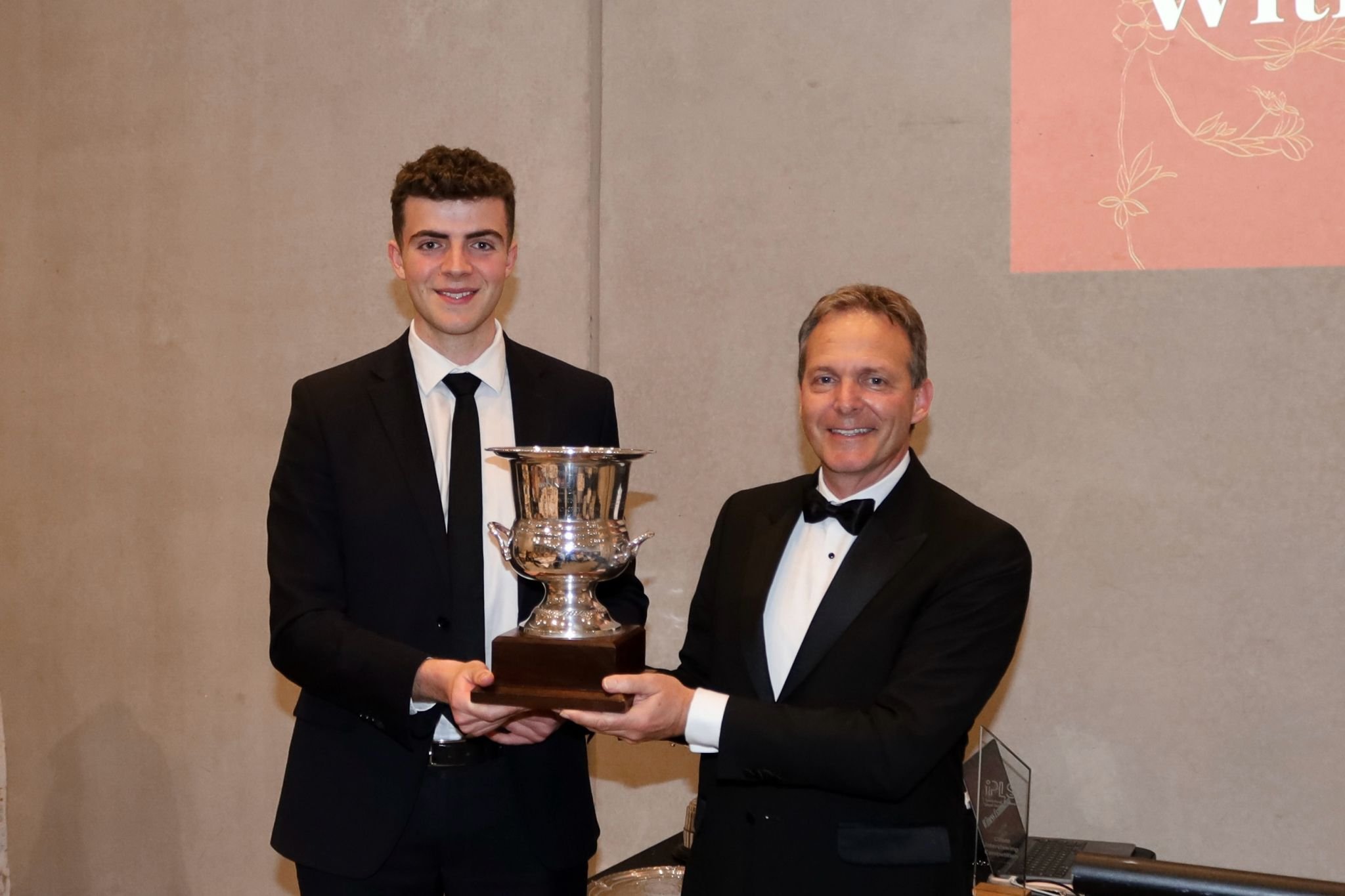 Two men in suits holding a silver trophy, smiling at an event.