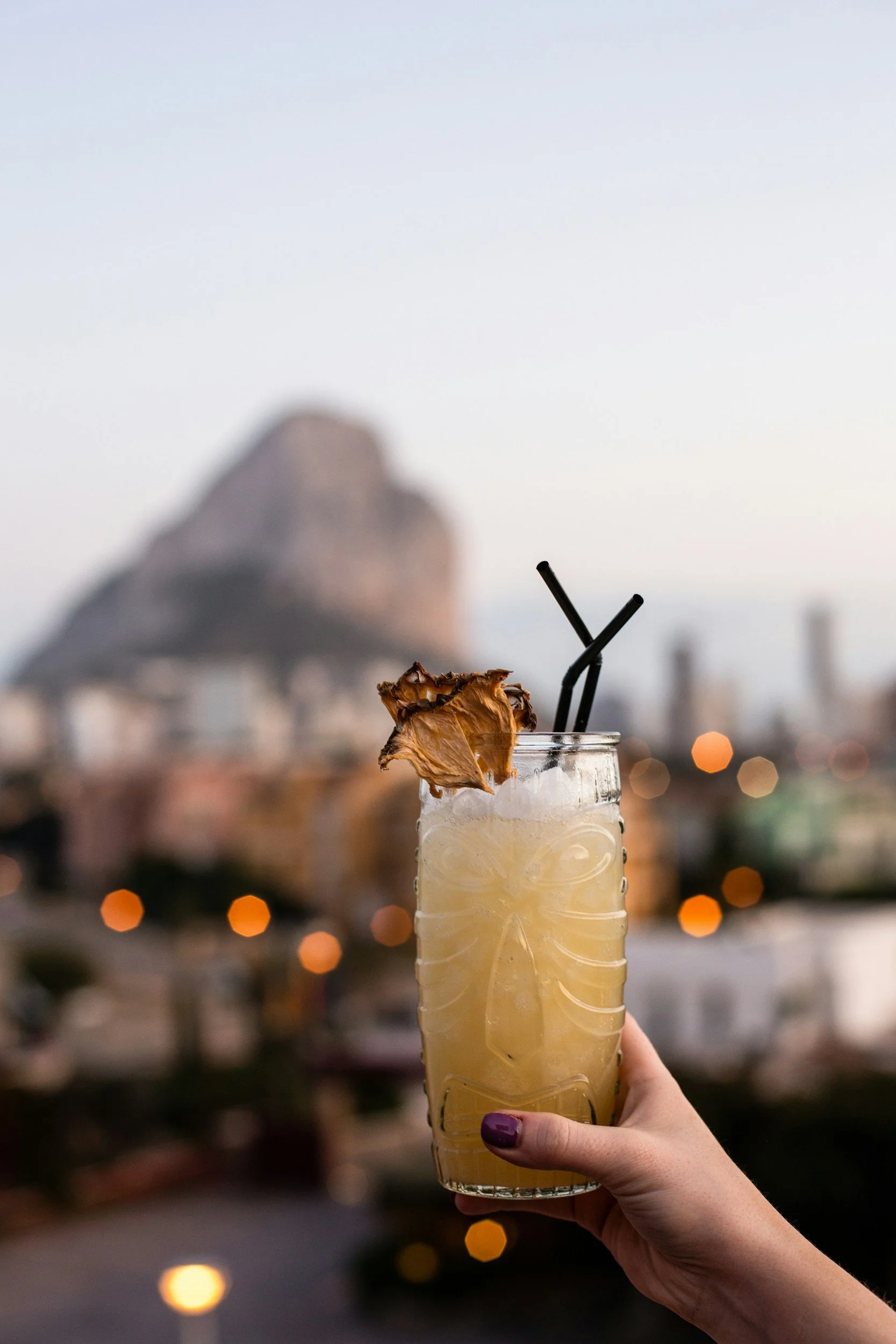 Un verre à cocktail tiki contenant une boisson jaune, décoré d'une tranche d'ananas séchée, avec deux pailles noires, tenu par une main avec ongles violets, avec une ville et une colline en arrière-plan lors du coucher du soleil.