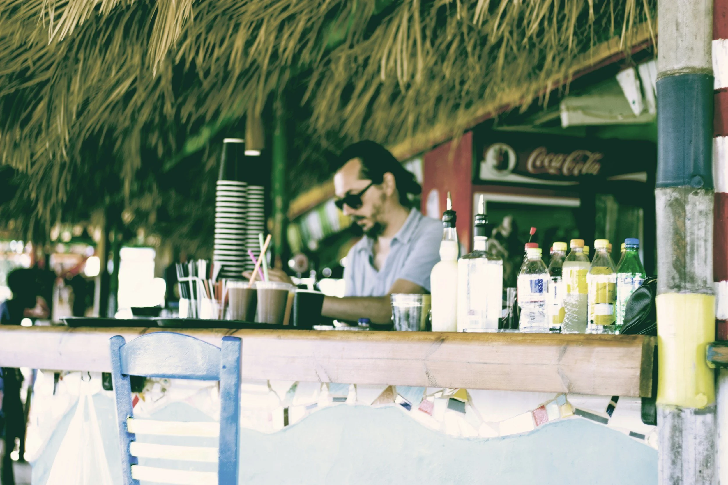 Un homme portant des lunettes de soleil et une chemise blanche travaille derrière un bar de plage avec des bouteilles et des gobelets, sous un toit en feuilles de palmier.