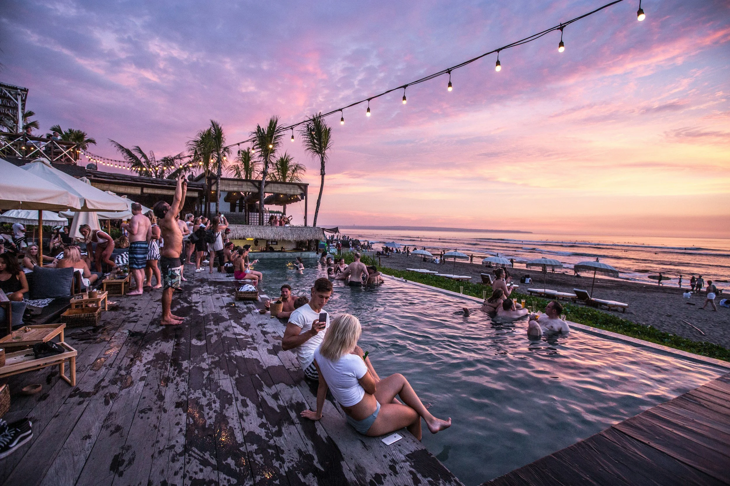 Scène de plage animée pendant le coucher de soleil avec des personnes dans une piscine et sur le sable, des parasols, des palmiers et un ciel coloré.