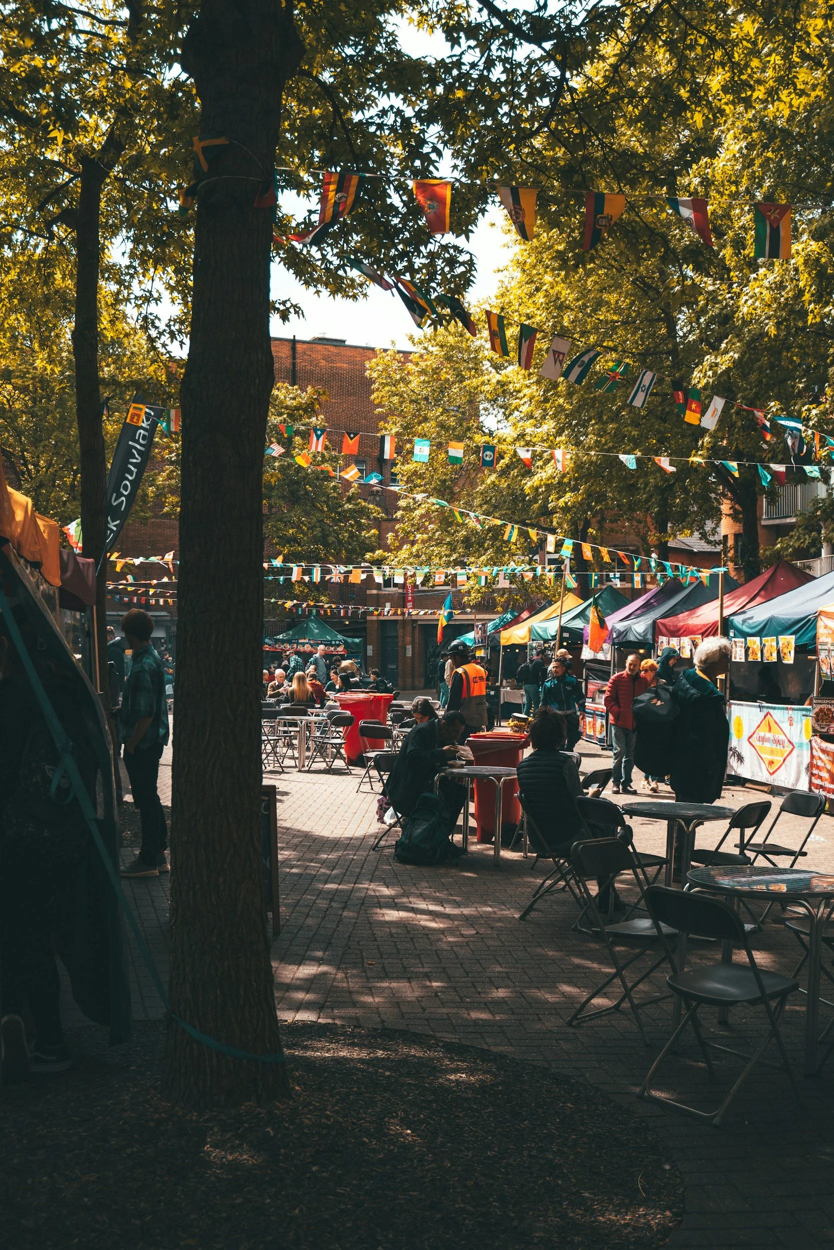 Marché en plein air avec des stands colorés, des personnes assises et discutant, des drapeaux de plusieurs pays suspendus entre les arbres, ambiance ensoleillée et ombragée.