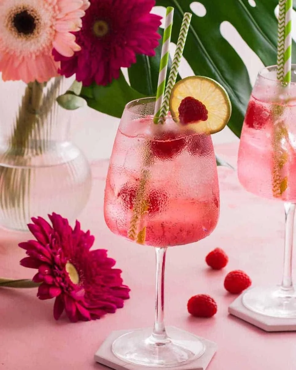 Deux verres de boisson rose avec des tranches de citron, des framboises et des pailles vertes et blanches, sur une table rose avec des fleurs et des framboises.