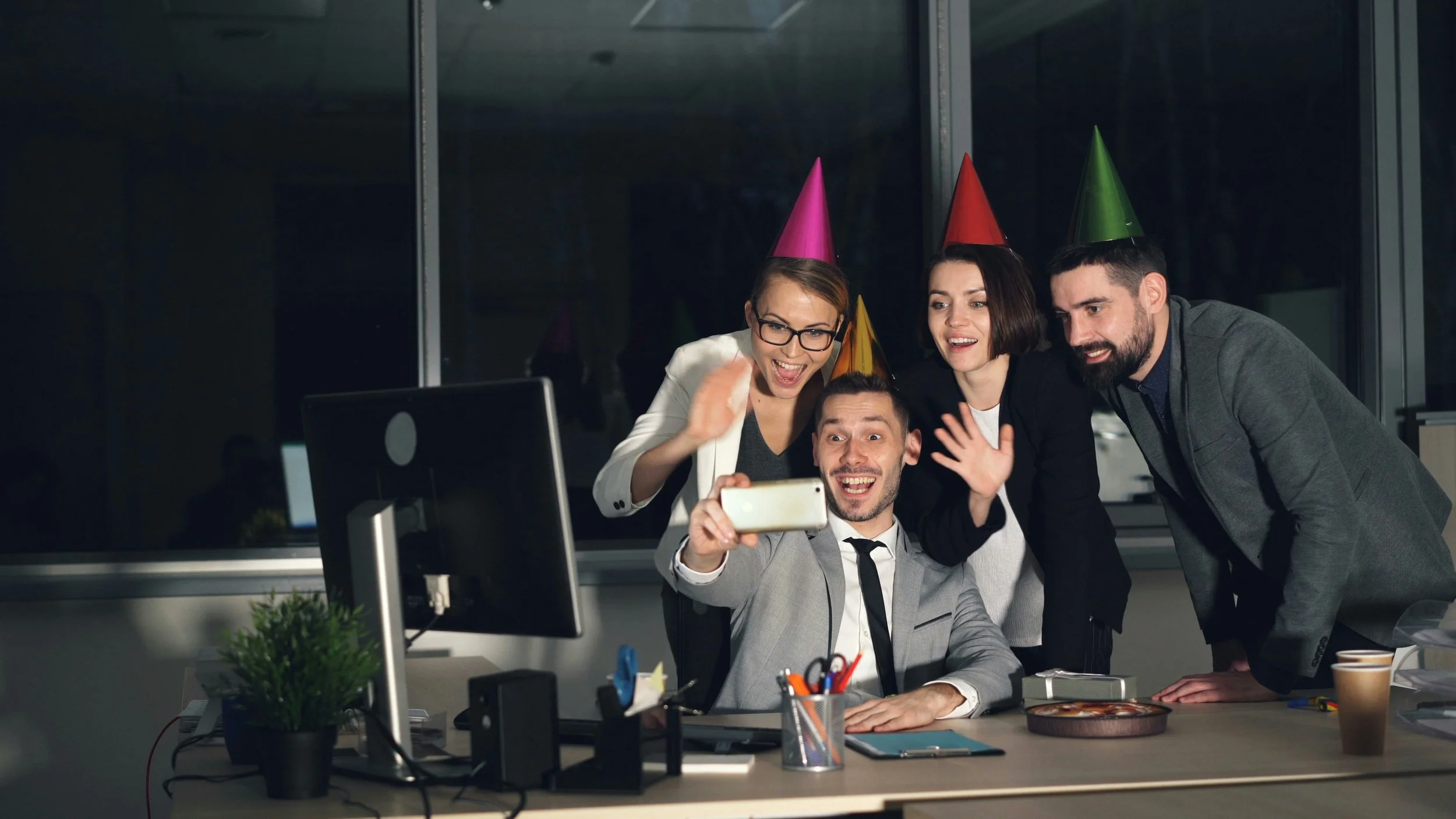 Groupe de quatre personnes au bureau portant des chapeaux de fête, souriant et prenant une photo d'eux-mêmes avec un smartphone, célébrant une occasion spéciale.
