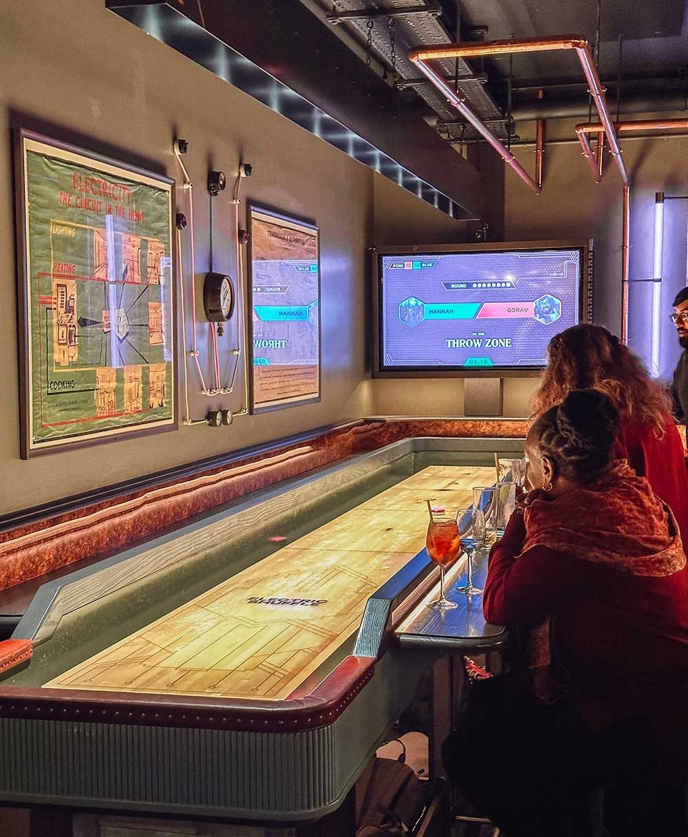 Un groupe de personnes jouent à un jeu de shuffleboard dans un bar avec un décor industriel, des écrans tactiles affichant des scores et un tableau illustrant le circuit électrique.