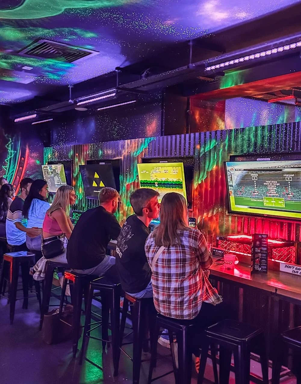 Des personnes jouent à des jeux vidéo sur des écrans dans un bar ou une salle de jeux avec un décor lumineux coloré et un ciel étoilé au plafond.