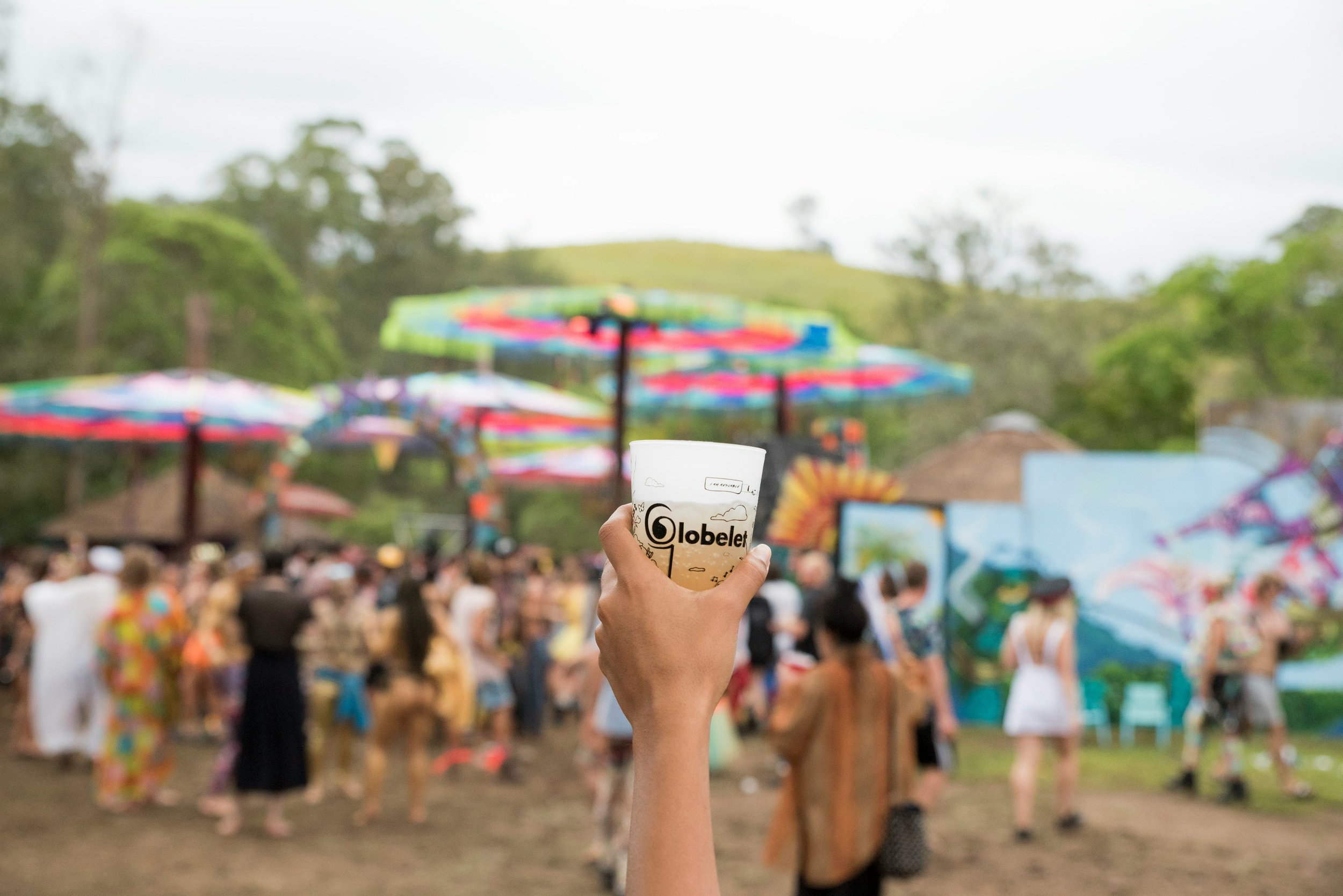 Une main tient un gobelet avec le mot "Gobelet" devant un festival en plein air avec des gens, des parasols colorés et un décor naturel verdoyant.