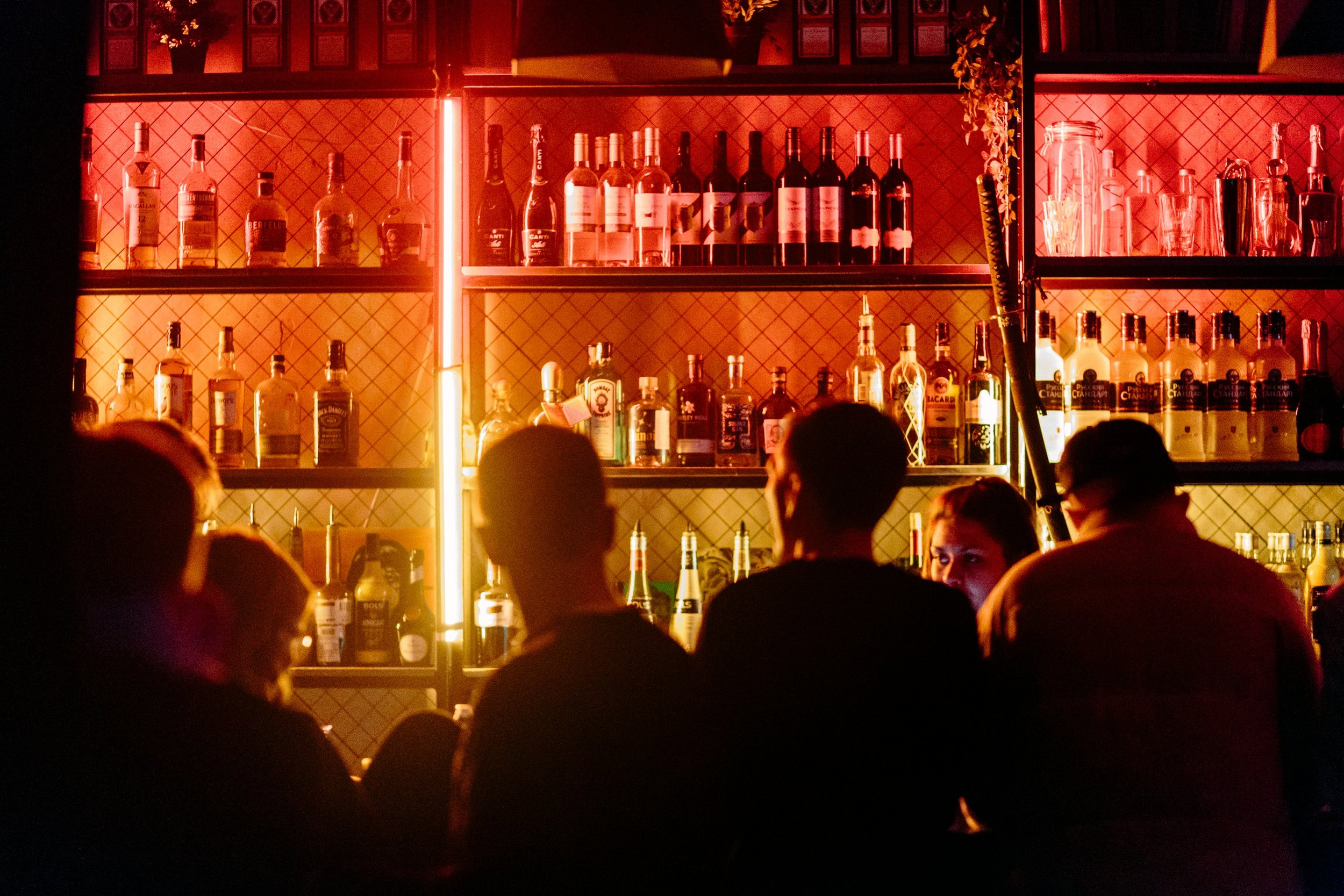 Un bar avec des étagères remplies de bouteilles de différentes boissons alcoolisées, éclairé par des lumières chaudes, et plusieurs personnes assises ou debout en conversation.