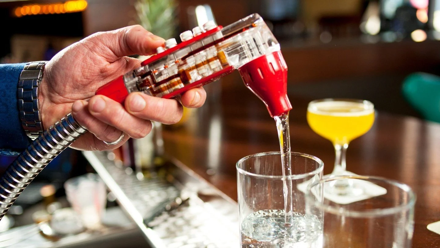 Une personne verse un mélange de cocktails dans un verre, avec d'autres cocktails colorés en arrière-plan.