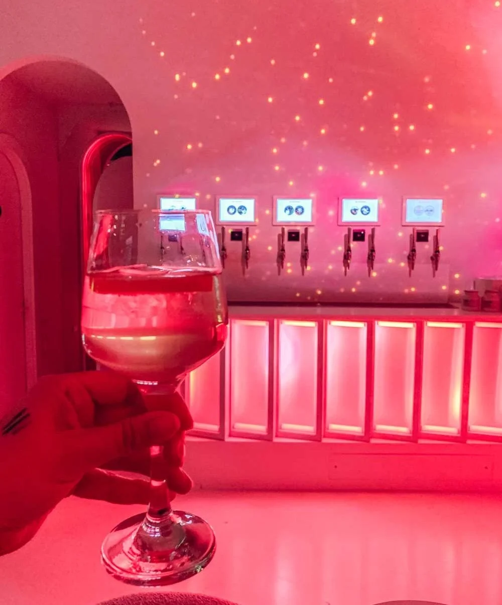 Verre de Hugo spritz par une main devant un mur rose avec des projections lumineuses et un distributeur de boissons à cinq robinets avec écrans lumineux au fond.