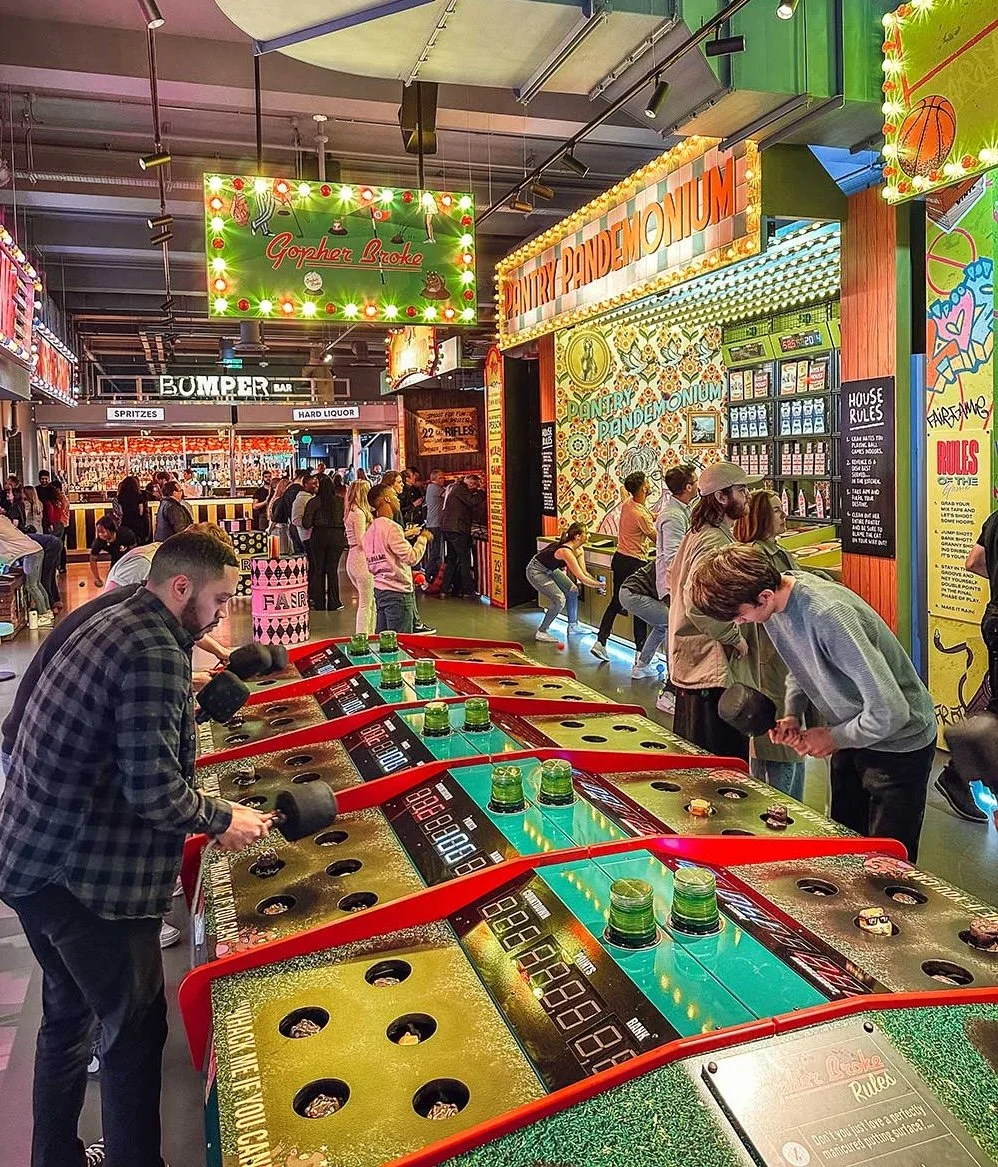 Un centre de divertissement intérieur avec plusieurs personnes jouant. Il y a des tables de jeux de palets et des zones d'amusement avec des enfants et des adultes, des lumières colorées, et divers panneaux lumineux.