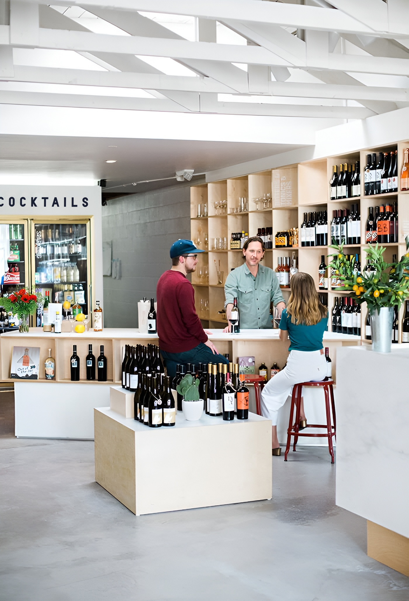 Un bar à vin moderne avec trois personnes : un homme derrière le comptoir, un homme avec une casquette bleue et une femme en blanc qui commandent du vin.