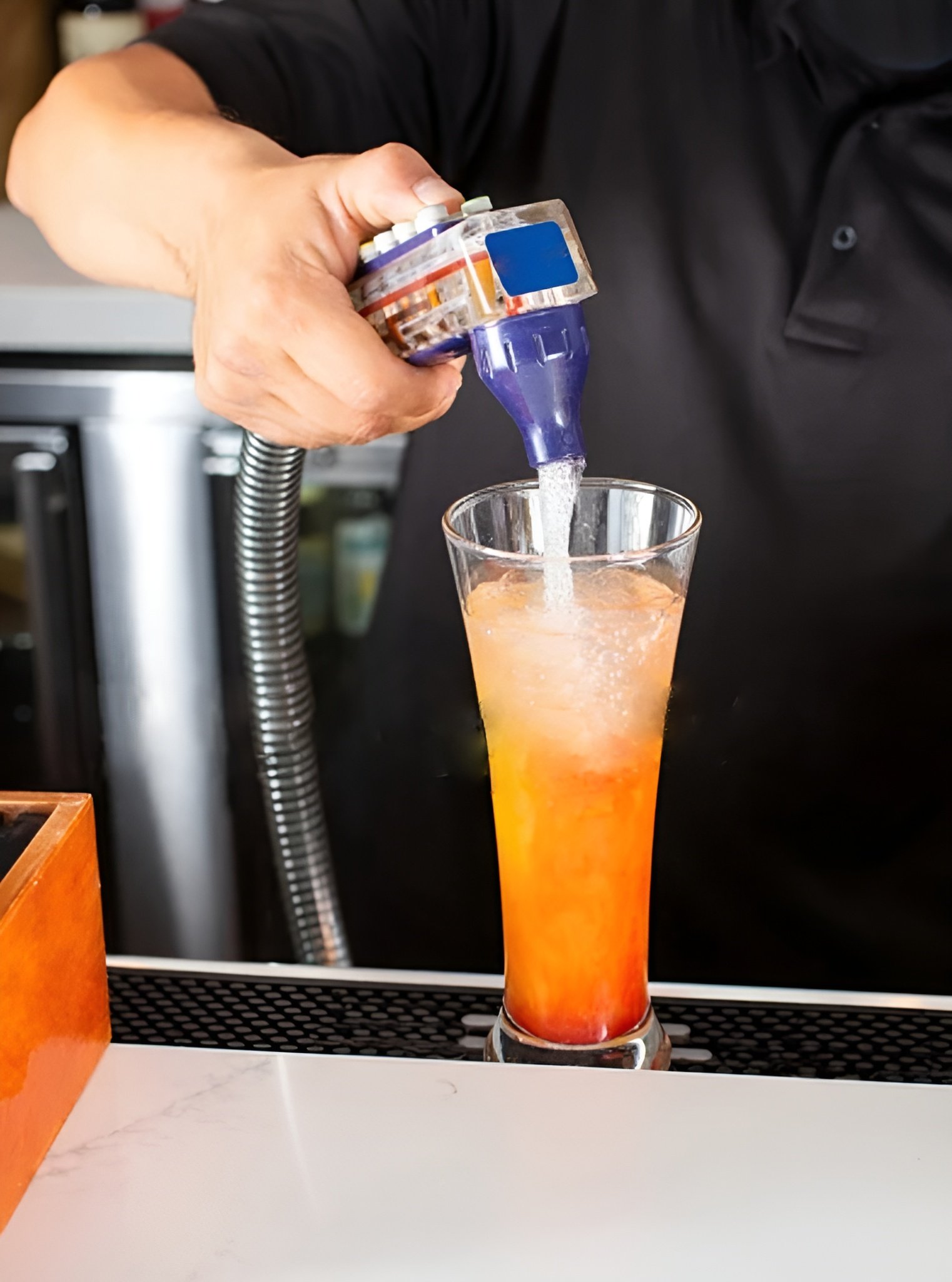 Une personne verse une boisson dans un verre haut en forme de flûte, contenant un cocktail coloré orange et rouge, à l'aide d'une bouteille en plastique. La personne porte une chemise noire et se trouve en arrière-plan d'un bar ou comptoir.
