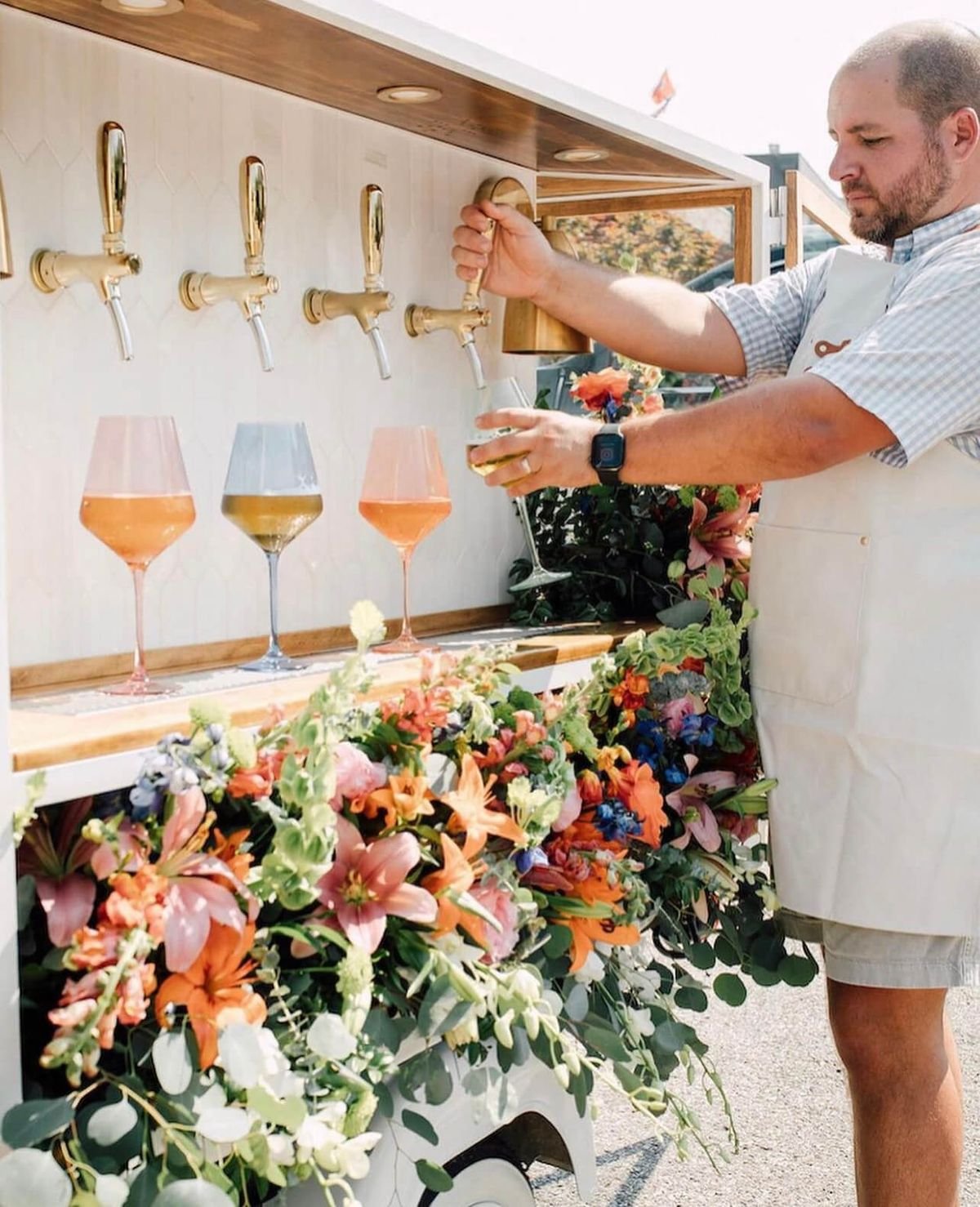 Un homme verse du vin dans un verre à partir d'un robinet, avec des fleurs colorées en dessous et trois autres verres avec différents vins sur un comptoir