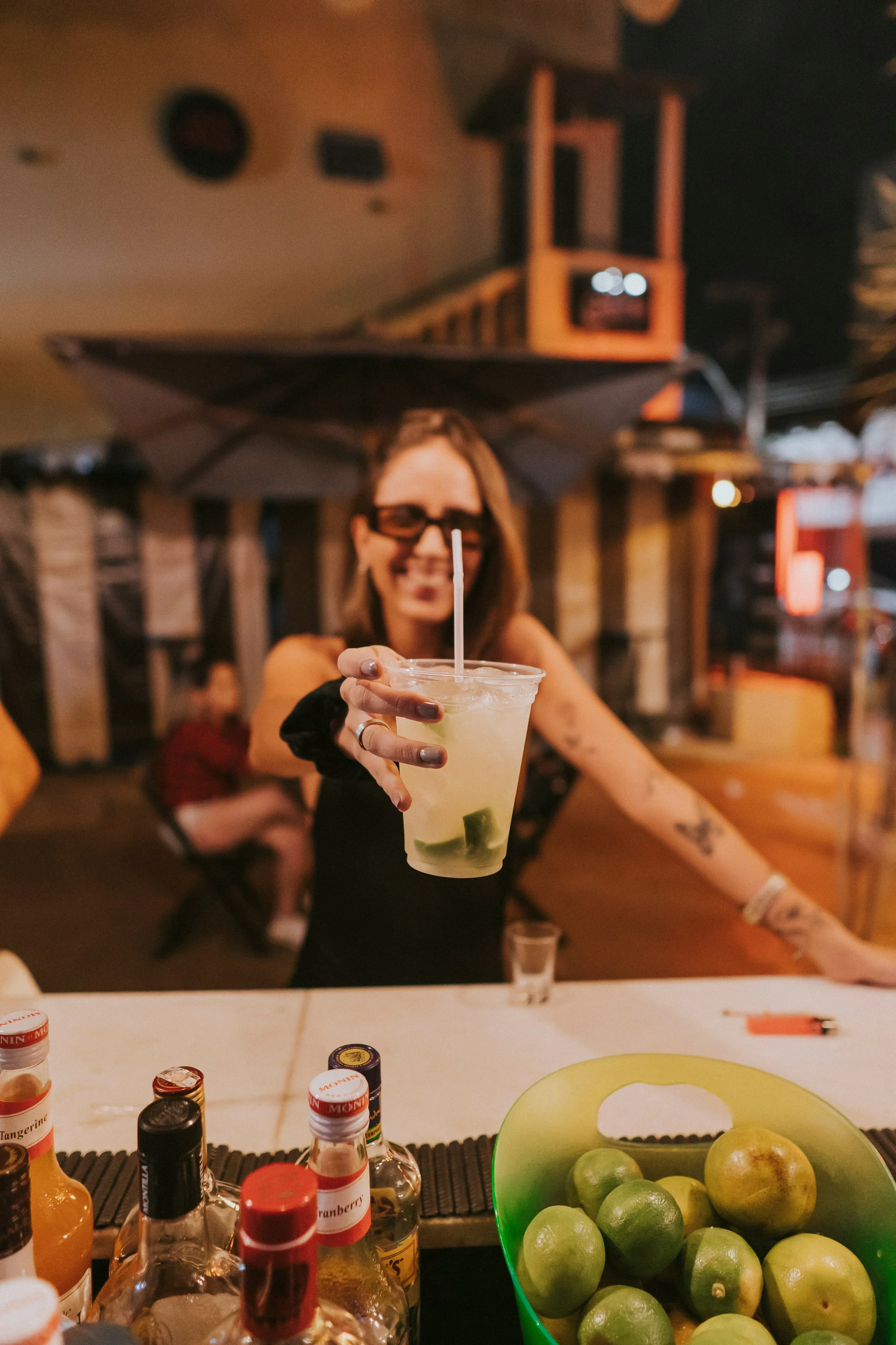 Une femme souriante portant des lunettes de soleil, tenant un cocktail avec des citrons dans un bar ou restaurant en extérieur, avec plusieurs bouteilles sur le comptoir.