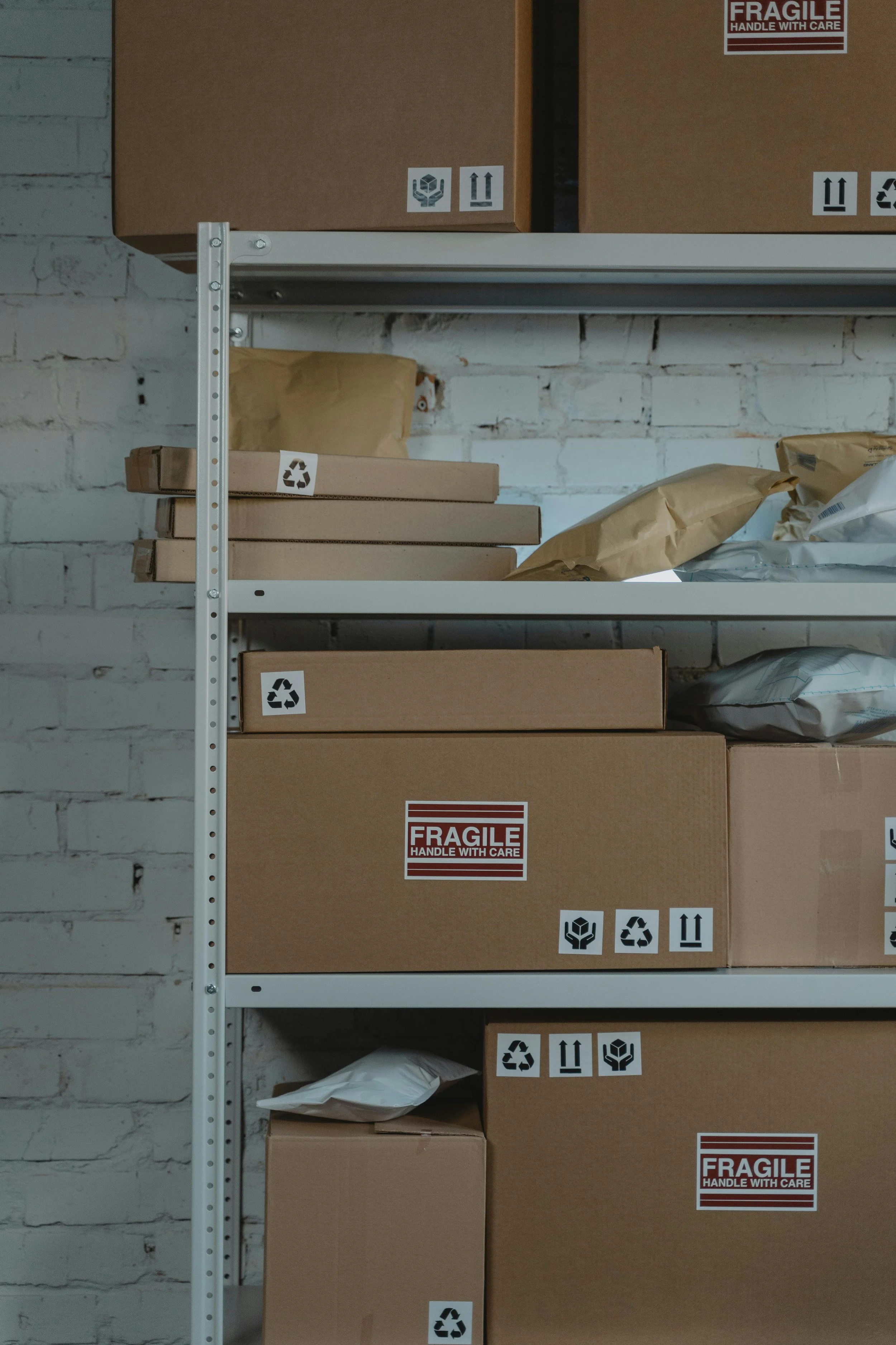 Shelves with cardboard boxes and packages, some labeled with 'FRAGILE handle with care' and recycling symbols, against a white brick wall.