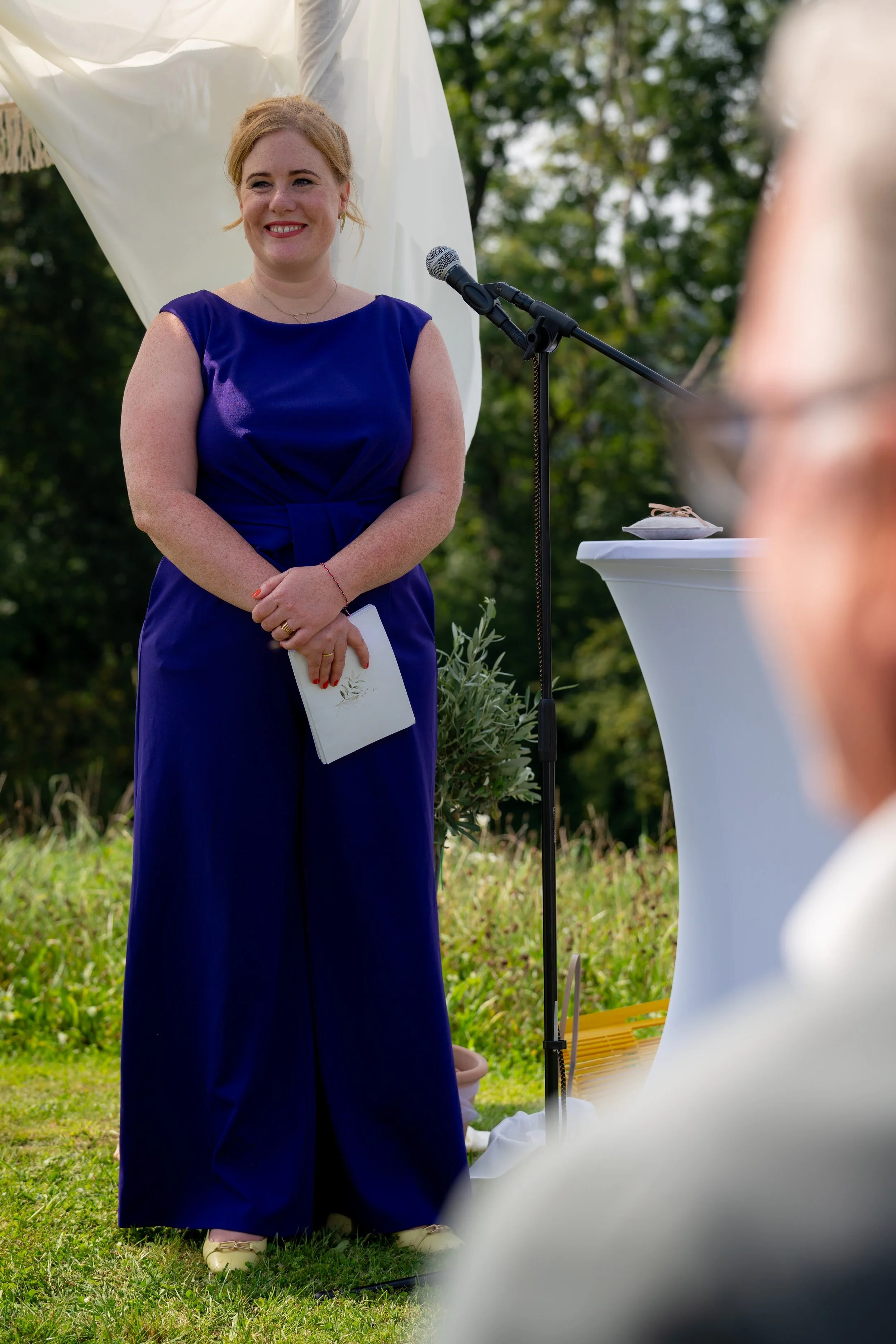 Eine Lächelnde Frau in einem blauen Kleid steht draußen bei einer Veranstaltung, neben einem Mikrofon und einem Stehtisch, im Hintergrund grüne Bäume.