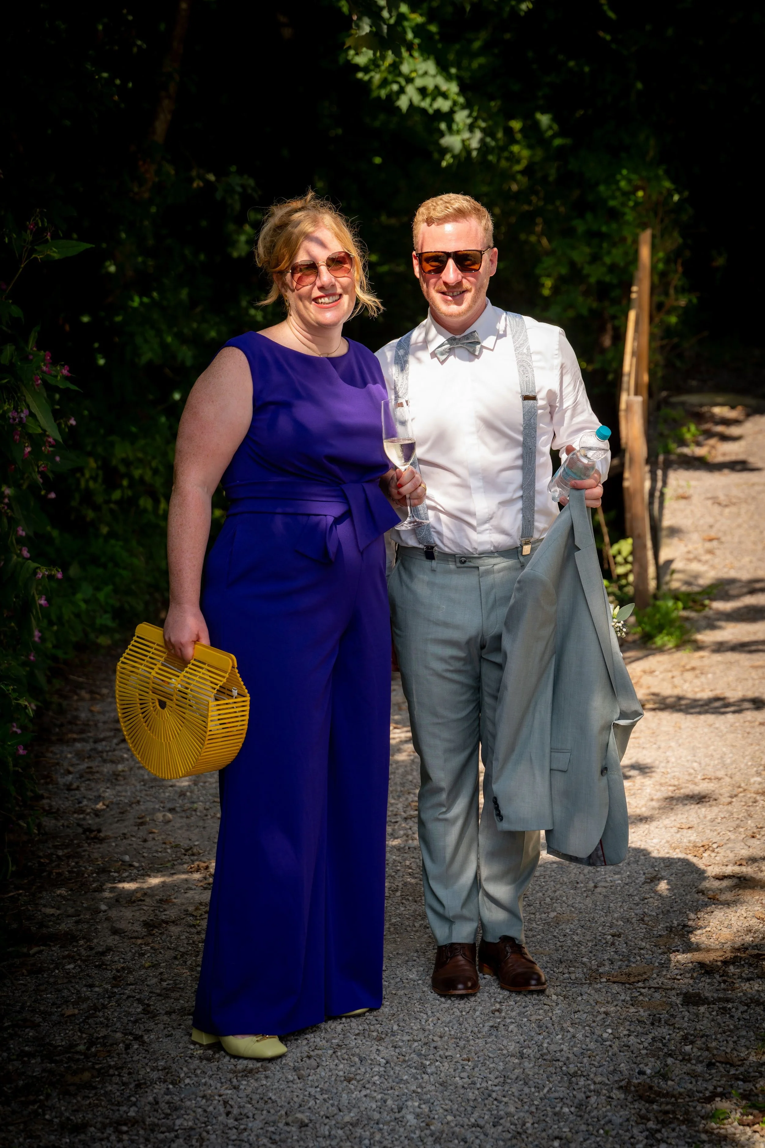 Ein lachendes Paar in formeller Kleidung bei sonnigem Wetter im Grünen, die Frau in einer blauen Abendrobe mit gelber Handtasche, der Mann in grauem Anzug mit Sonnenbrille, beide mit Getränken in der Hand.