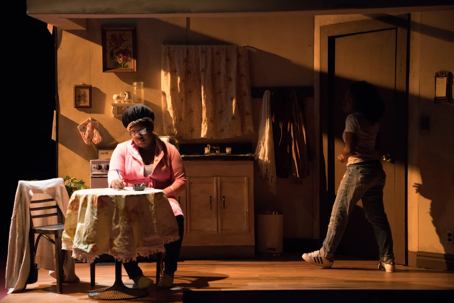 Stage scene of two women in a vintage-style kitchen; one woman seated at a dining table eating, wearing glasses and a pink cardigan, and another woman standing near a door, facing away, in casual clothing.