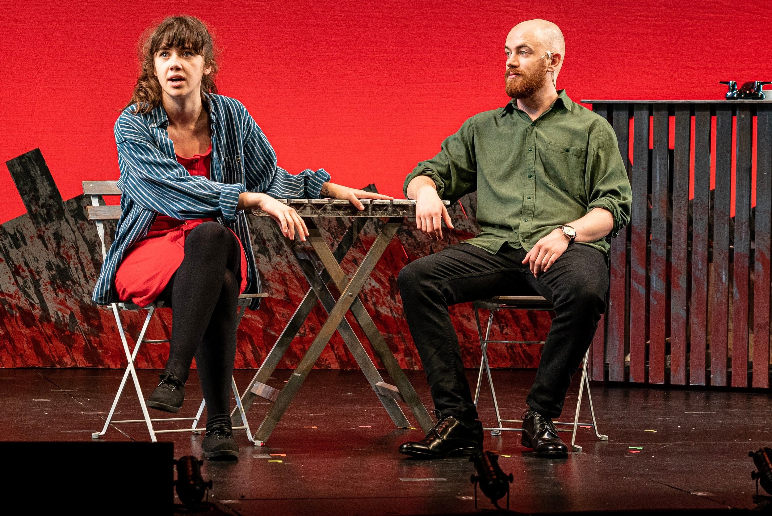 A woman with brown hair and a man with a beard sitting at a table on a stage, with a red background.