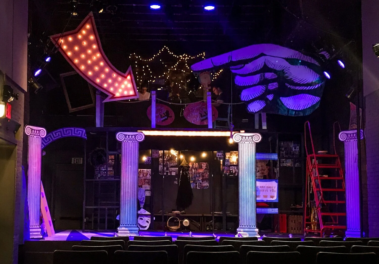 Theatre stage with Greek-style columns, purple lighting, a large pointing hand sign, a downward arrow sign with lights, and red stacks of chairs.