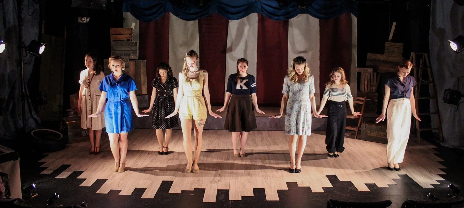 Nine women performing on stage, lined up and holding hands in a dance or theatrical production, with stage lighting and set decorations.