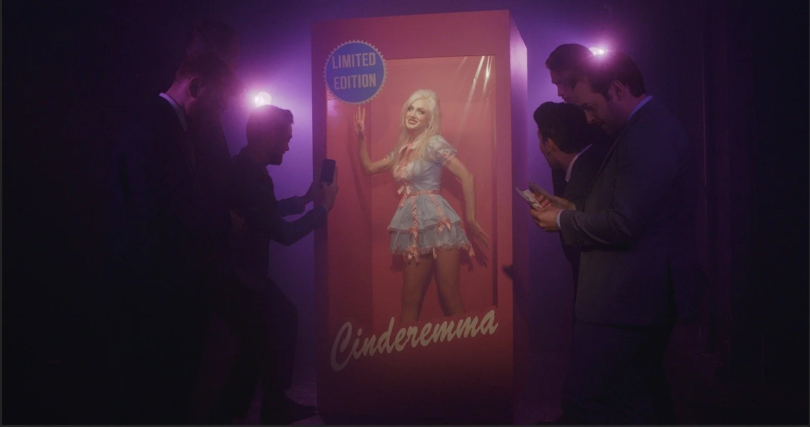 A woman in light blue dress with pink ribbons inside a pink display case marked 'Limited Edition,' surrounded by four men in suits taking photos or looking at their phones, with purple lighting.