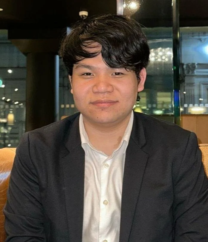 A young man with black hair, wearing a white dress shirt and black blazer, sitting indoors with a lit-up background and dark interior.