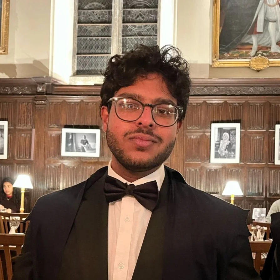 A young man with glasses and dark curly hair wearing a tuxedo with a bow tie, sitting in a fancy restaurant with wood-paneled walls and framed black-and-white photographs, and warm lighting from table lamps.