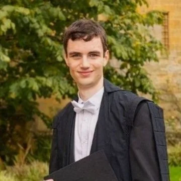 Young man in graduation cap and gown holding a diploma outdoors with trees and a building in the background.