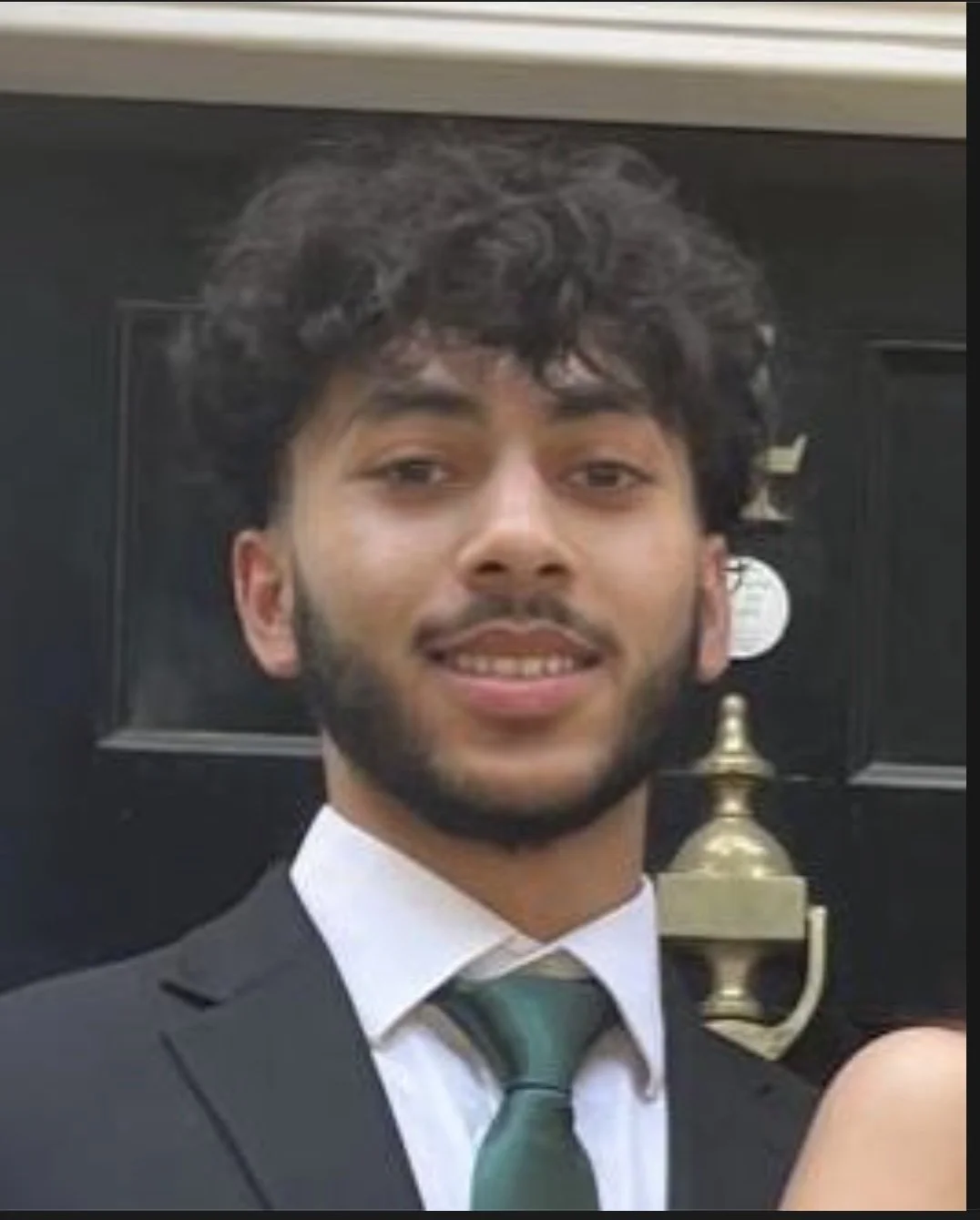 Young man with dark, curly hair and beard, dressed in a suit and tie, smiling, standing in front of a black door with a brass door knocker.