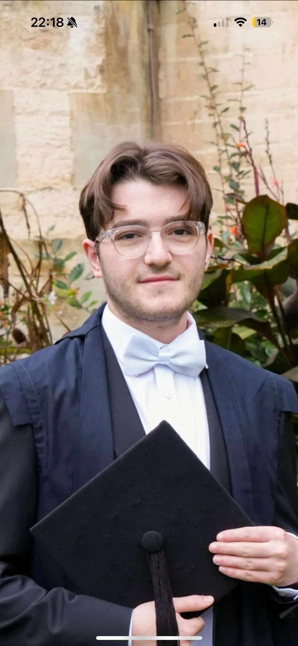 Young man in graduation gown and cap holding diploma outside near a brick wall and plants.