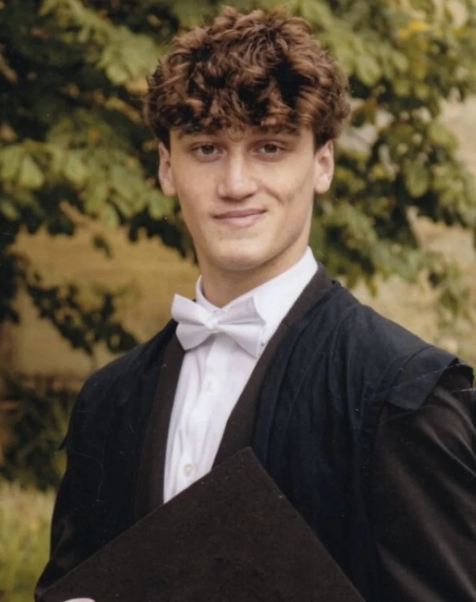 A young man in a graduation gown and white bow tie, holding a diploma, smiling outdoors with trees in the background.
