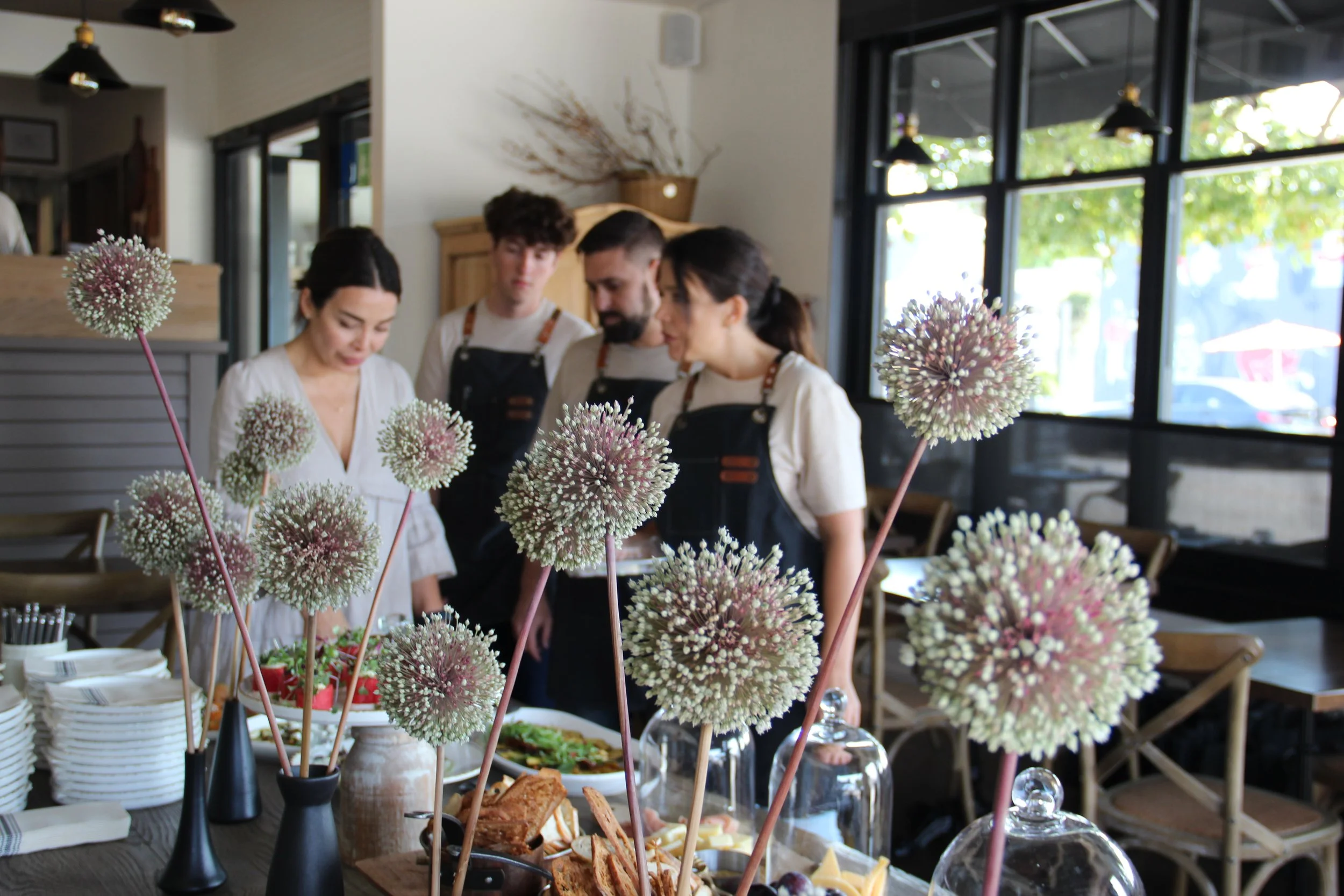 Group of four people in aprons gathered around a table with food and flowers in a restaurant with large windows.