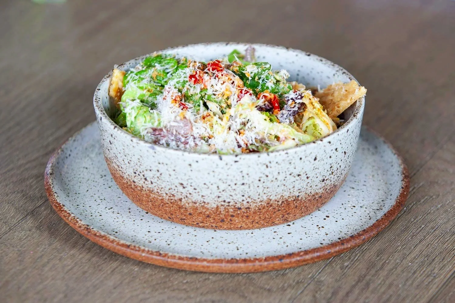 A bowl of Caesar salad with lettuce, grated cheese, croutons, and cherry tomatoes on a speckled plate placed on a wooden table.
