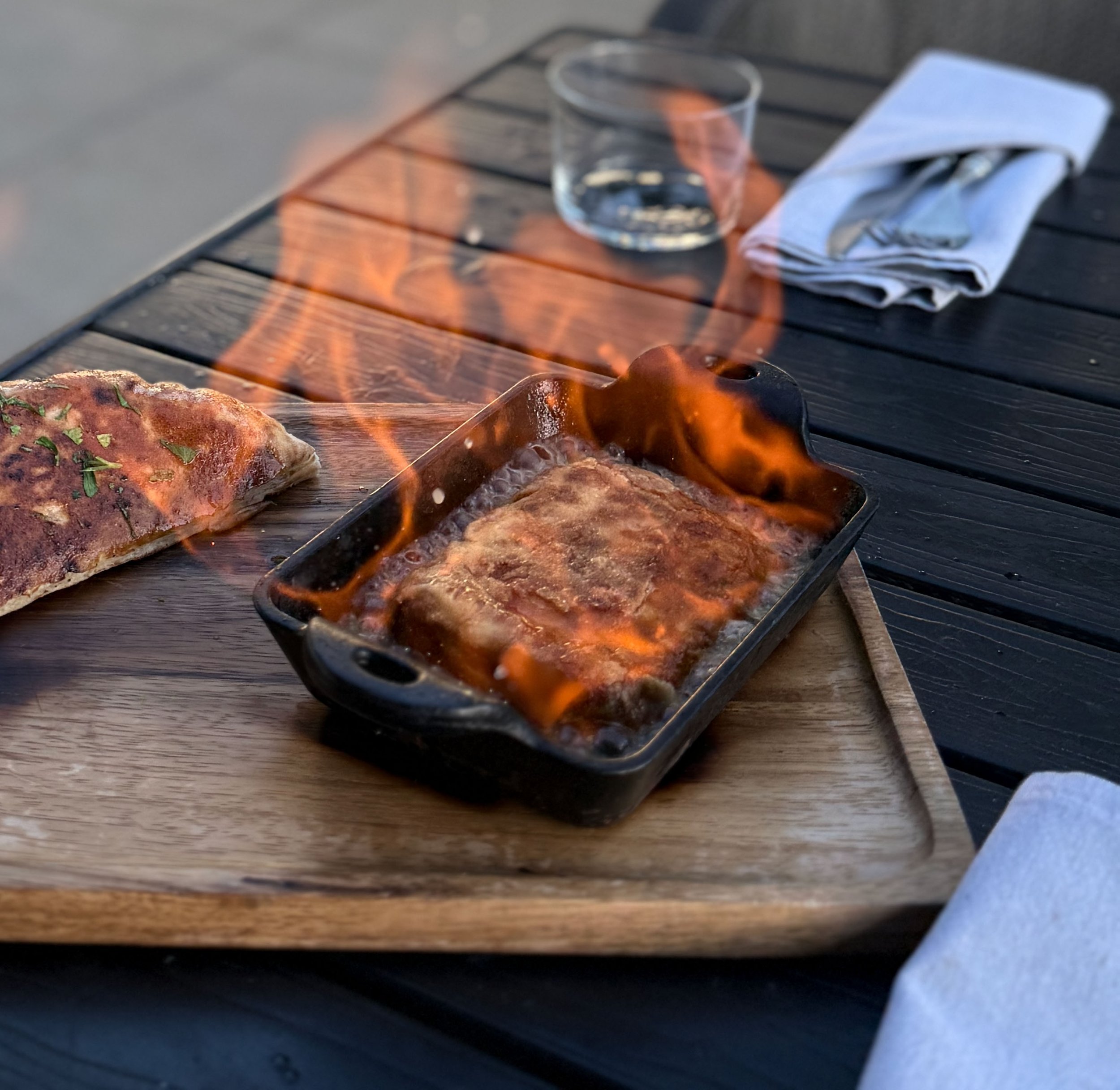 Grilled meat on a skillet with a glass of water, a napkin, and silverware on a napkin on a table.