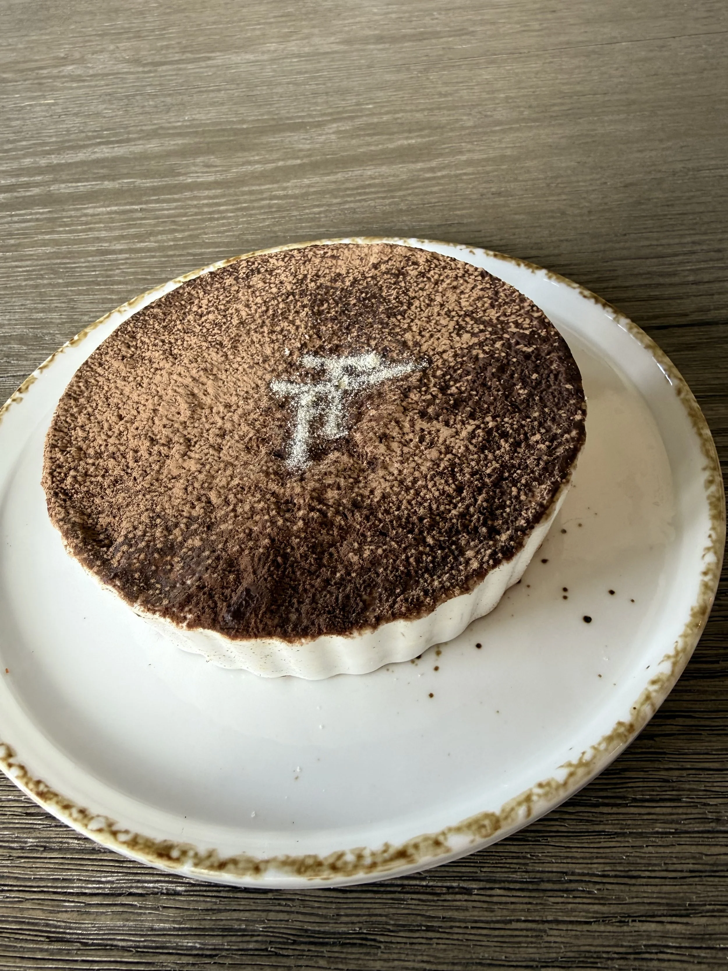 Close-up of a tiramisu dessert on a white plate with a brown rustic rim, placed on a wooden surface.