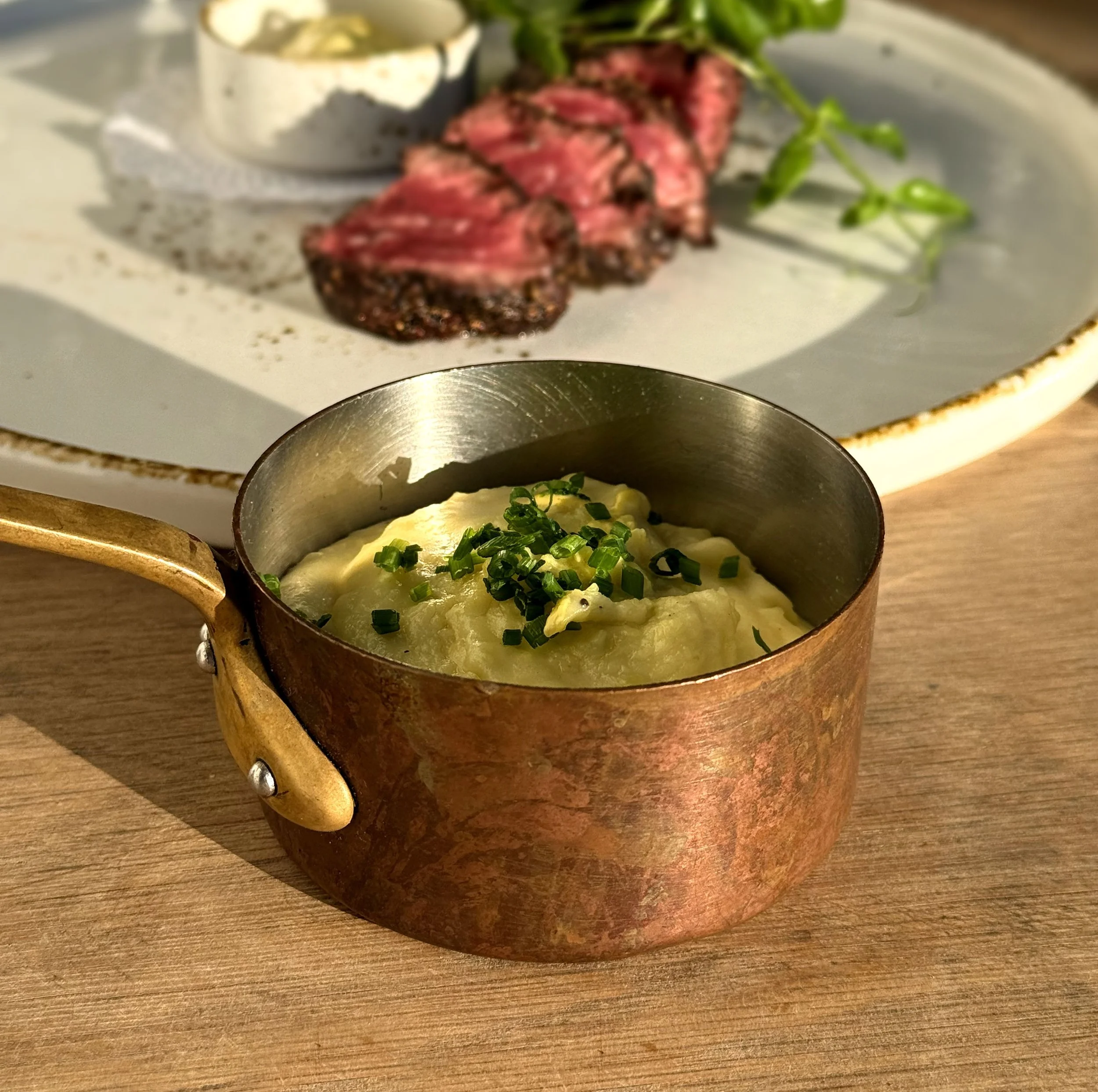 Sliced medium-rare steak with a pink center, garnished with chopped green onions on a white plate, with a creamy sauce in a small metallic bowl and a sprig of green herbs in the background.