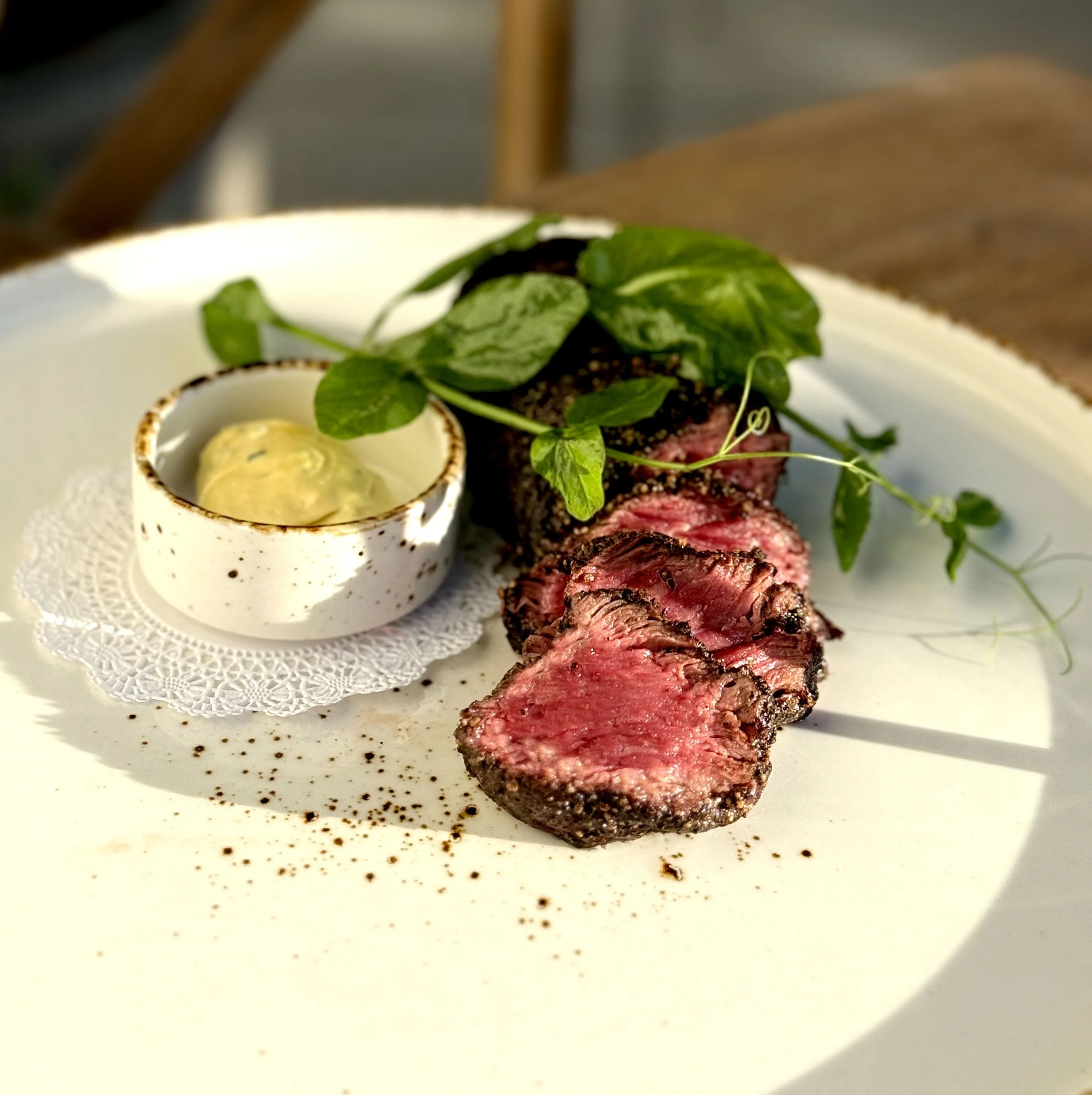 Sliced grilled steak on a white plate with a small cup of dipping sauce and fresh greens.