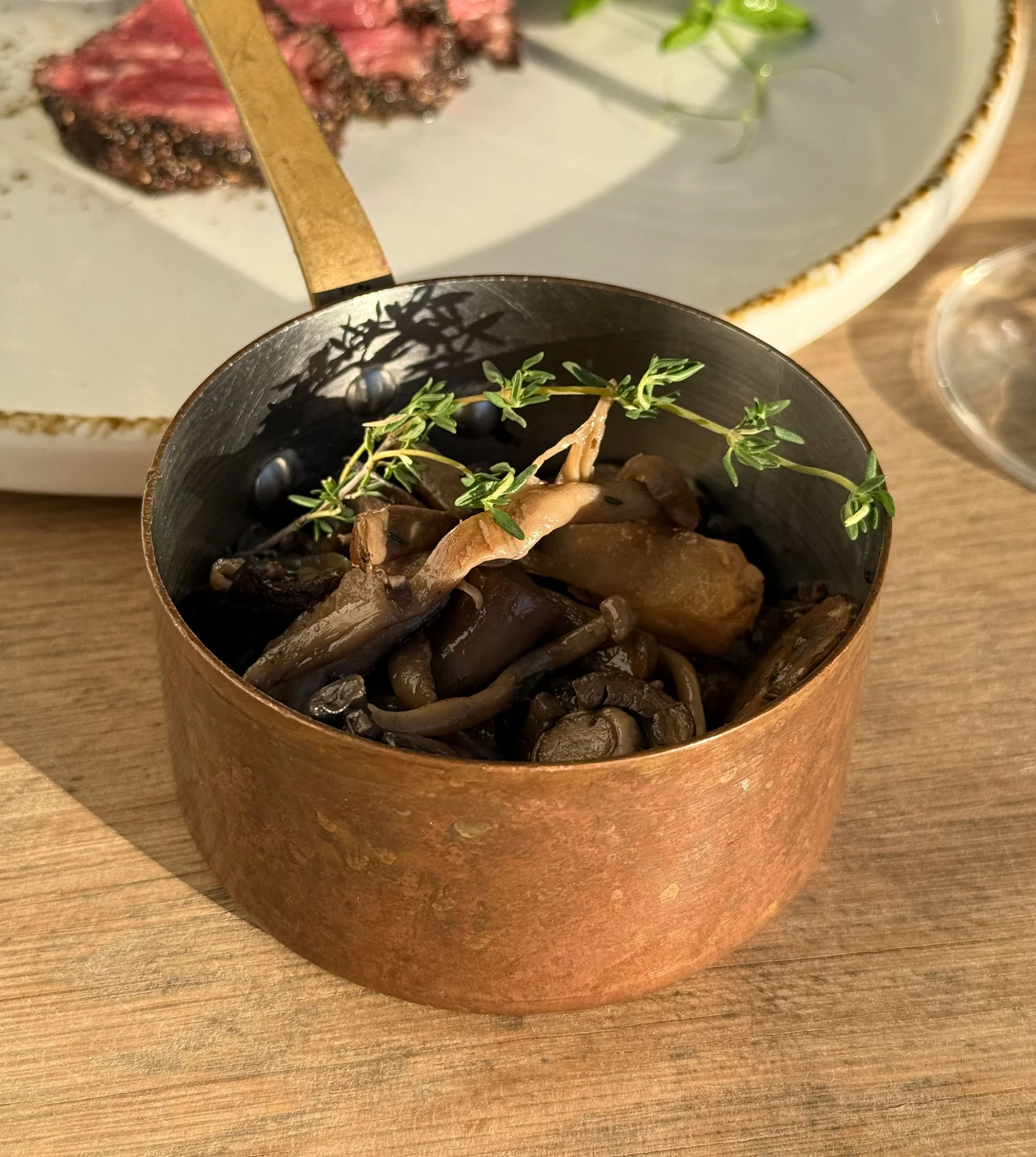 A close-up view of a copper-colored metal bowl with a rustic finish containing a plant with small green leaves, sitting on a wooden table. In the background, a ceramic dish with a white interior and brown rim is visible, holding a piece of cooked meat and some greens.