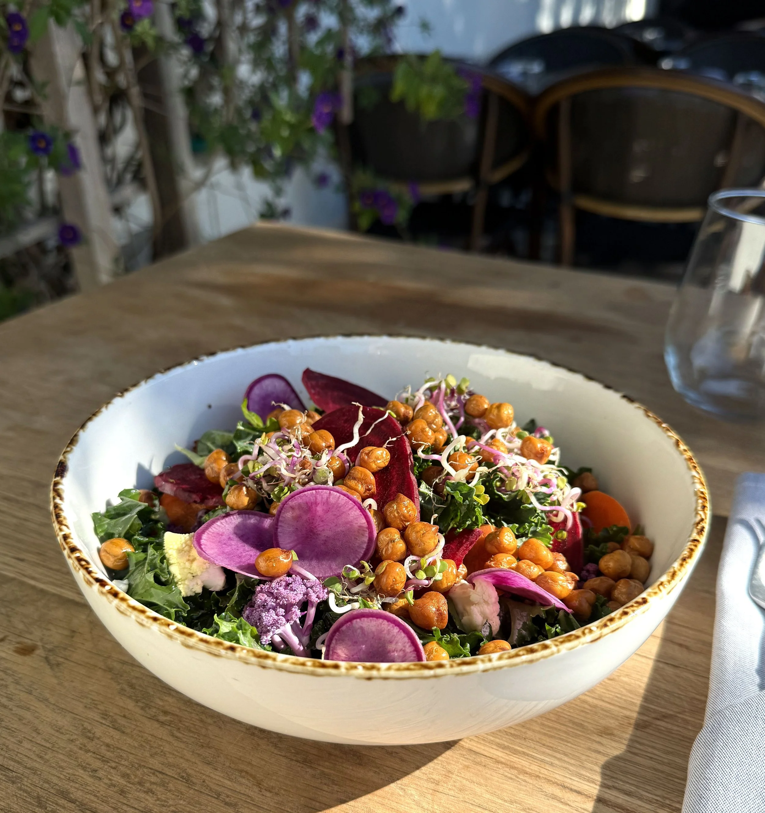 A white bowl with a decorative gold rim filled with a colorful vegetable salad, on a wooden table. The salad contains sliced purple radishes, chickpeas, sprouts, green vegetables, and purple edible flowers.