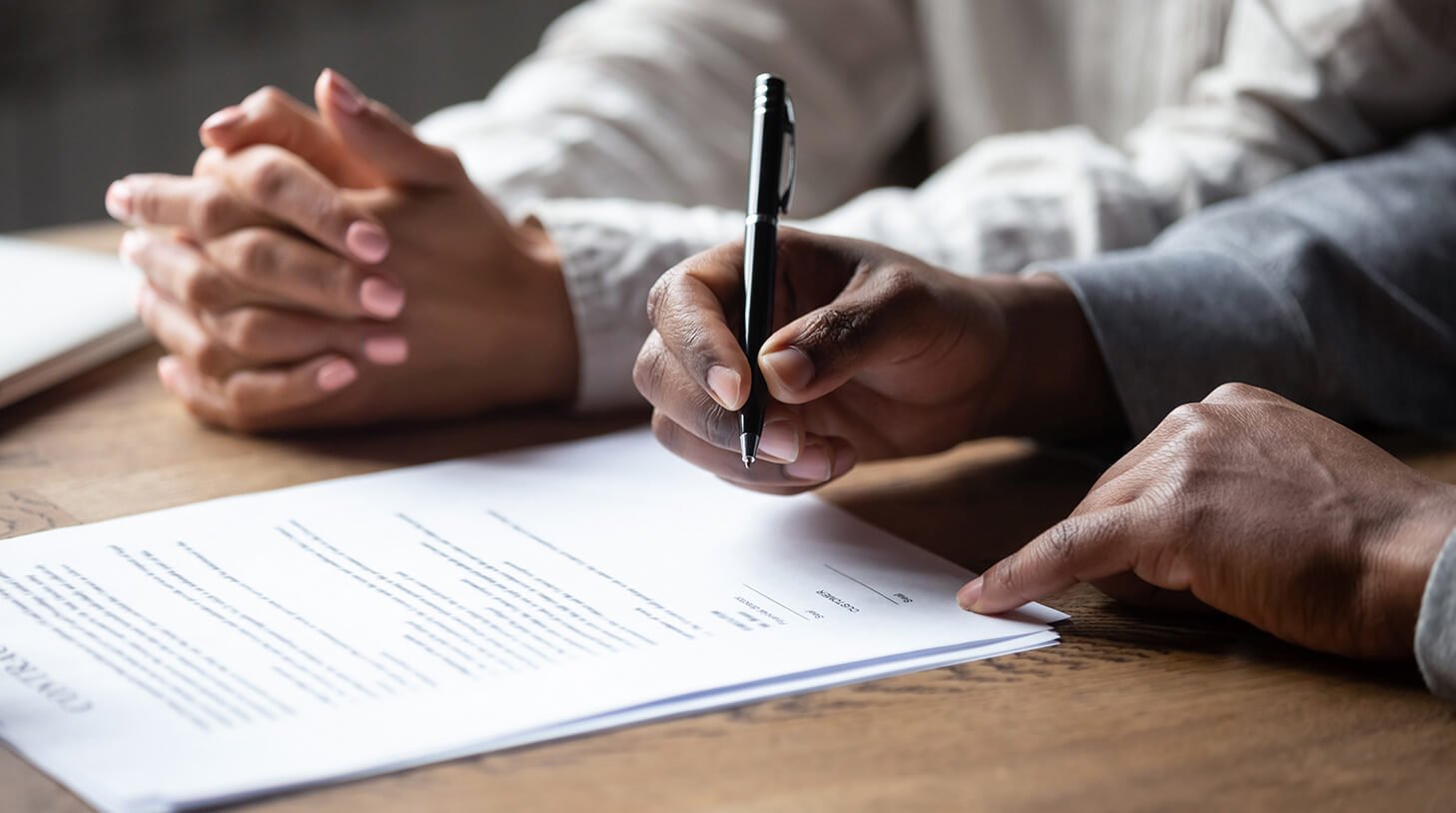 Two people sitting at a wooden table with documents, one is holding a pen and pointing at the paper, the other has hands clasped together.
