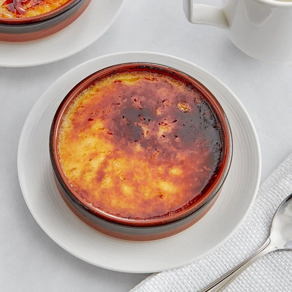 Crème brûlée with a caramelized sugar top served in a glass dish on a white plate, with a spoon and a cup of white sauce or cream nearby.
