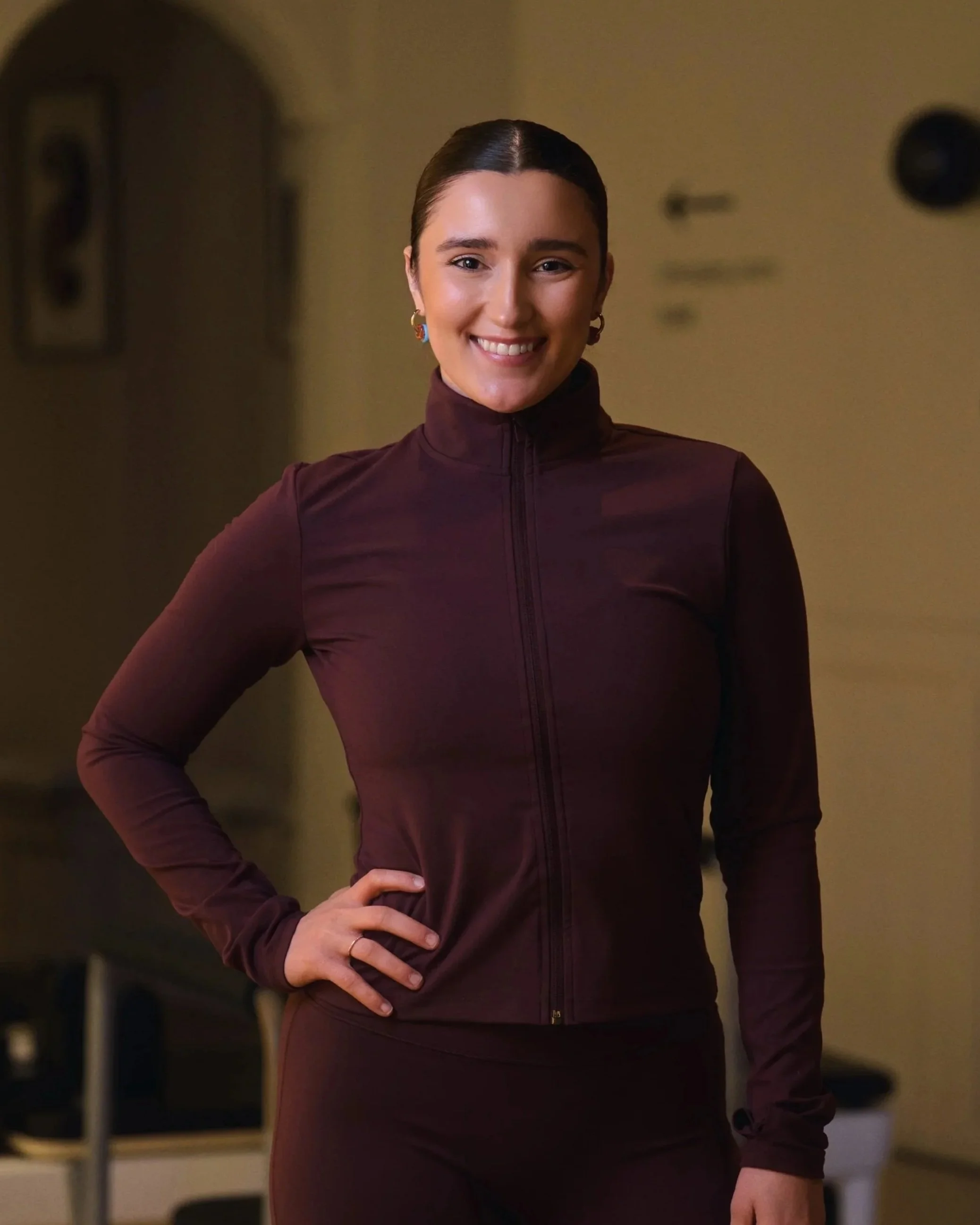 English speaking reformer instructor, posing in burgundy active wear, smiling in a beige reformer pilates studio